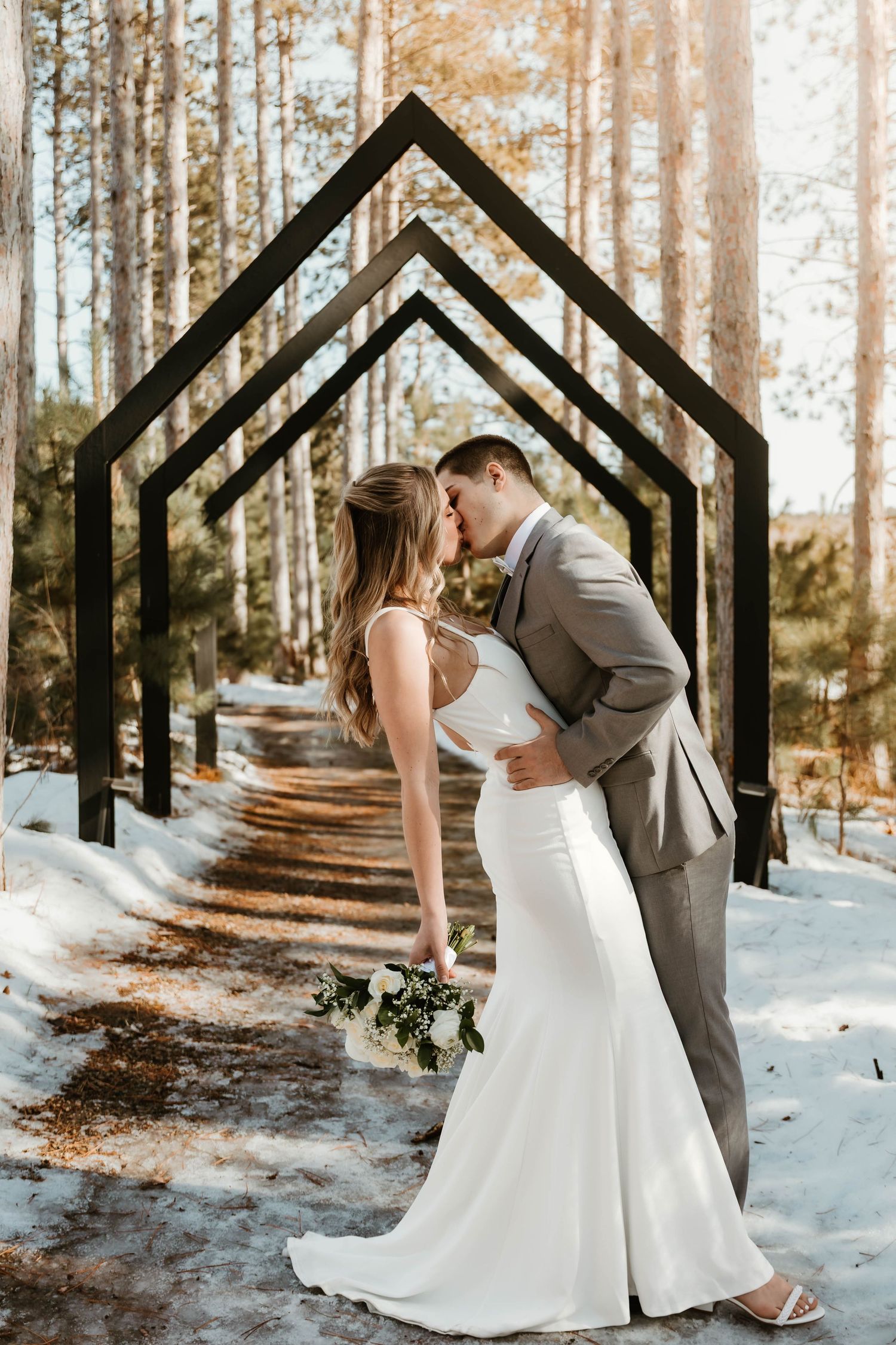 A romantic winter wedding photo series showing a couple kissing in a snowy forest with geometric wooden archways.