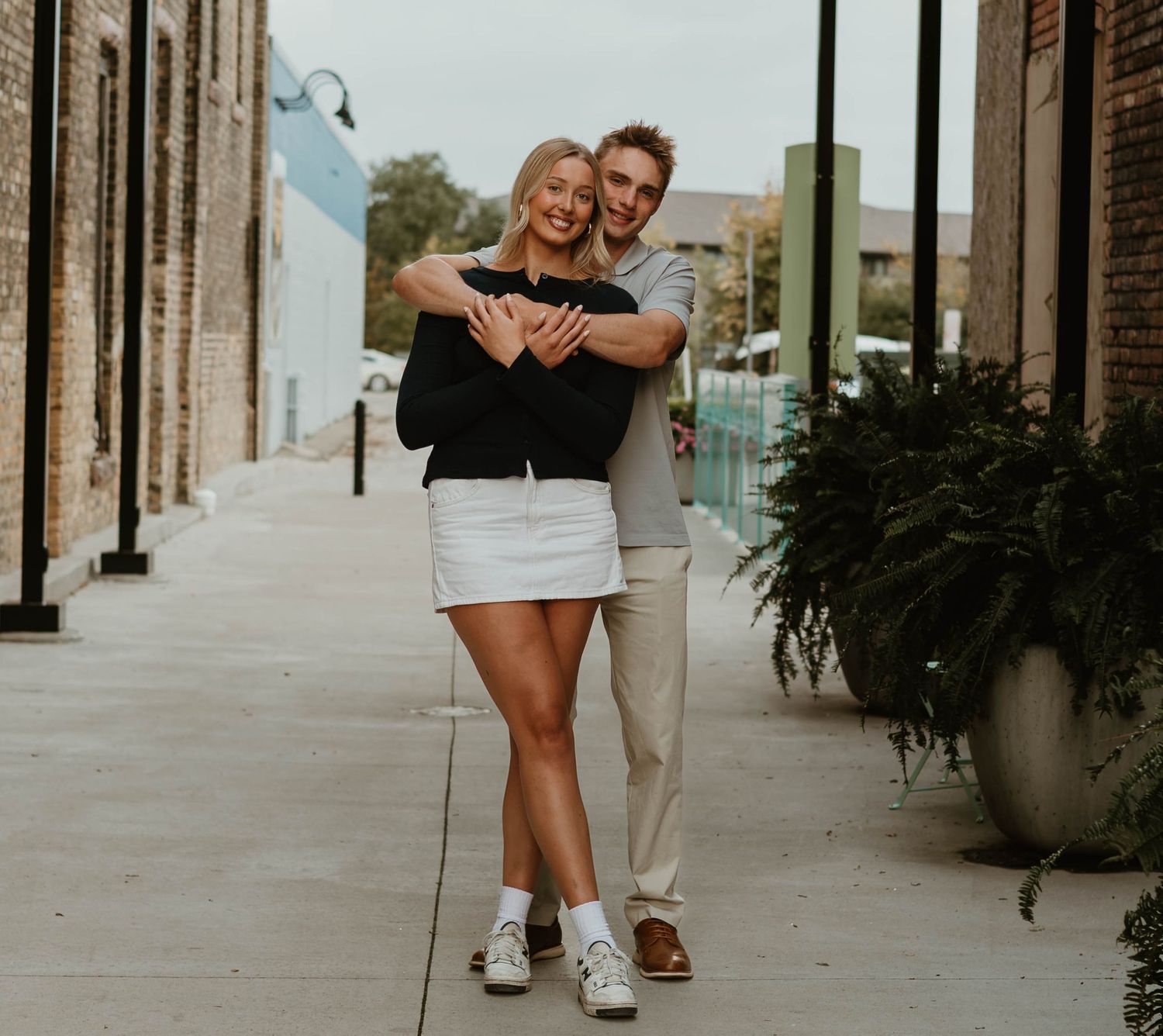 A couple embraces while walking down a sidewalk lined with brick buildings and decorative planters.