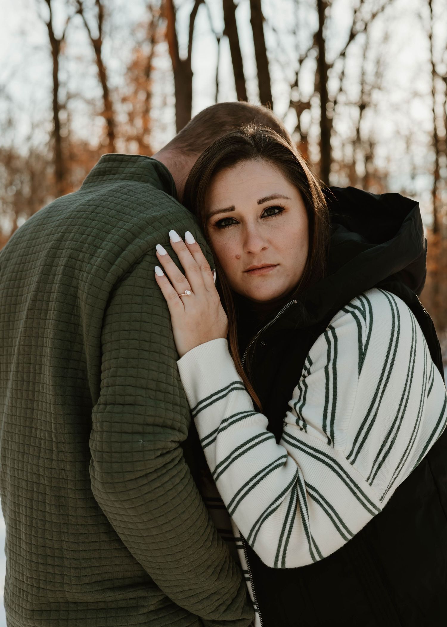 A couple in cozy sweaters shares an intimate moment while standing among bare trees on a winter day.