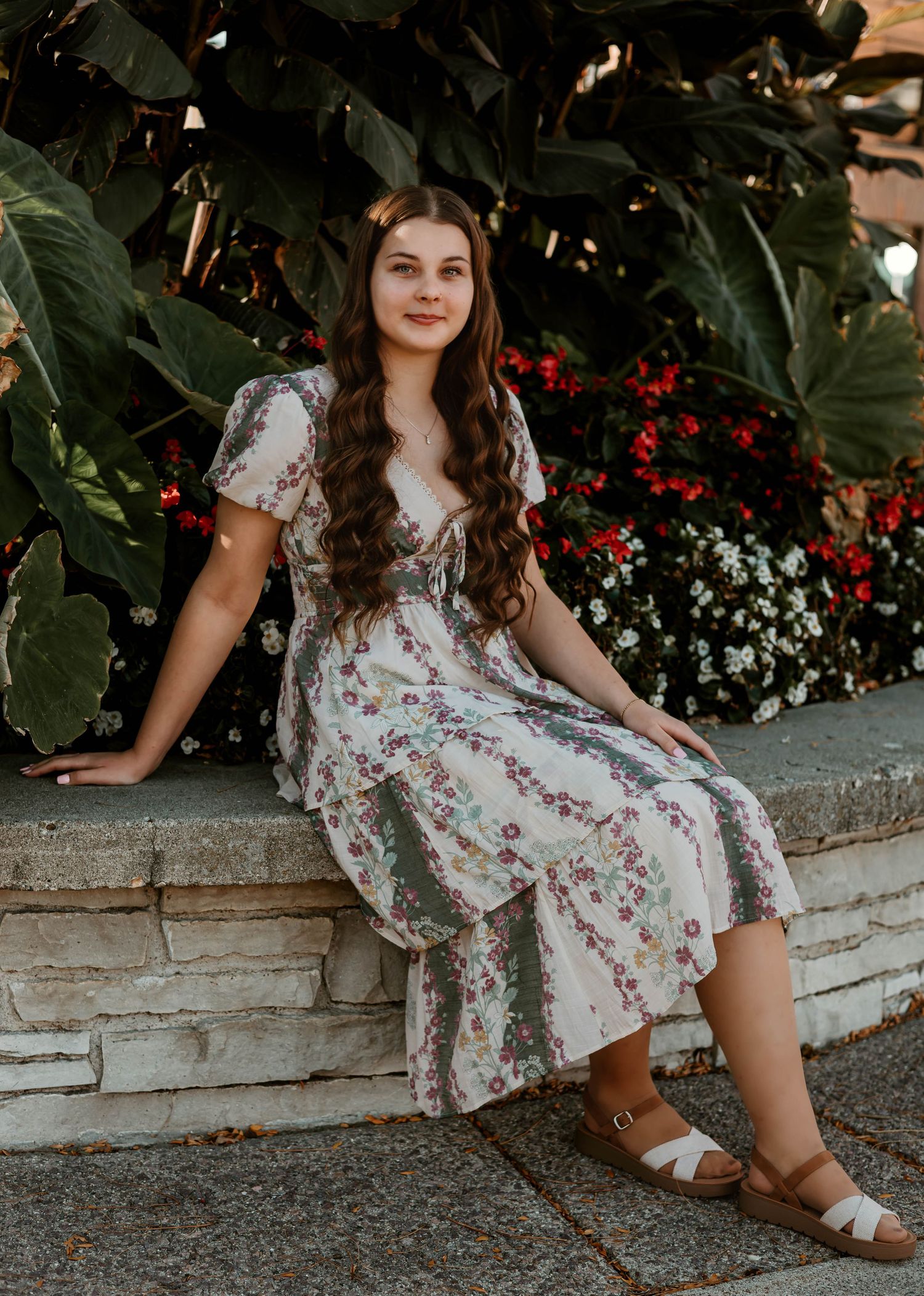 Young woman in floral midi dress sitting on stone ledge with lush greenery and red flowers behind her.