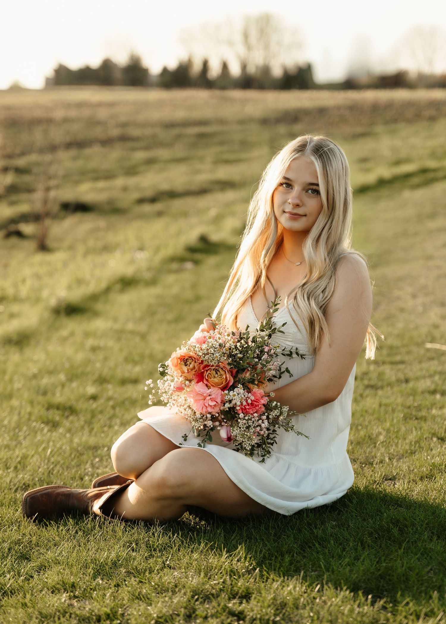 Young blonde woman in white dress holding pink floral bouquet, sitting on green grass field at golden hour.