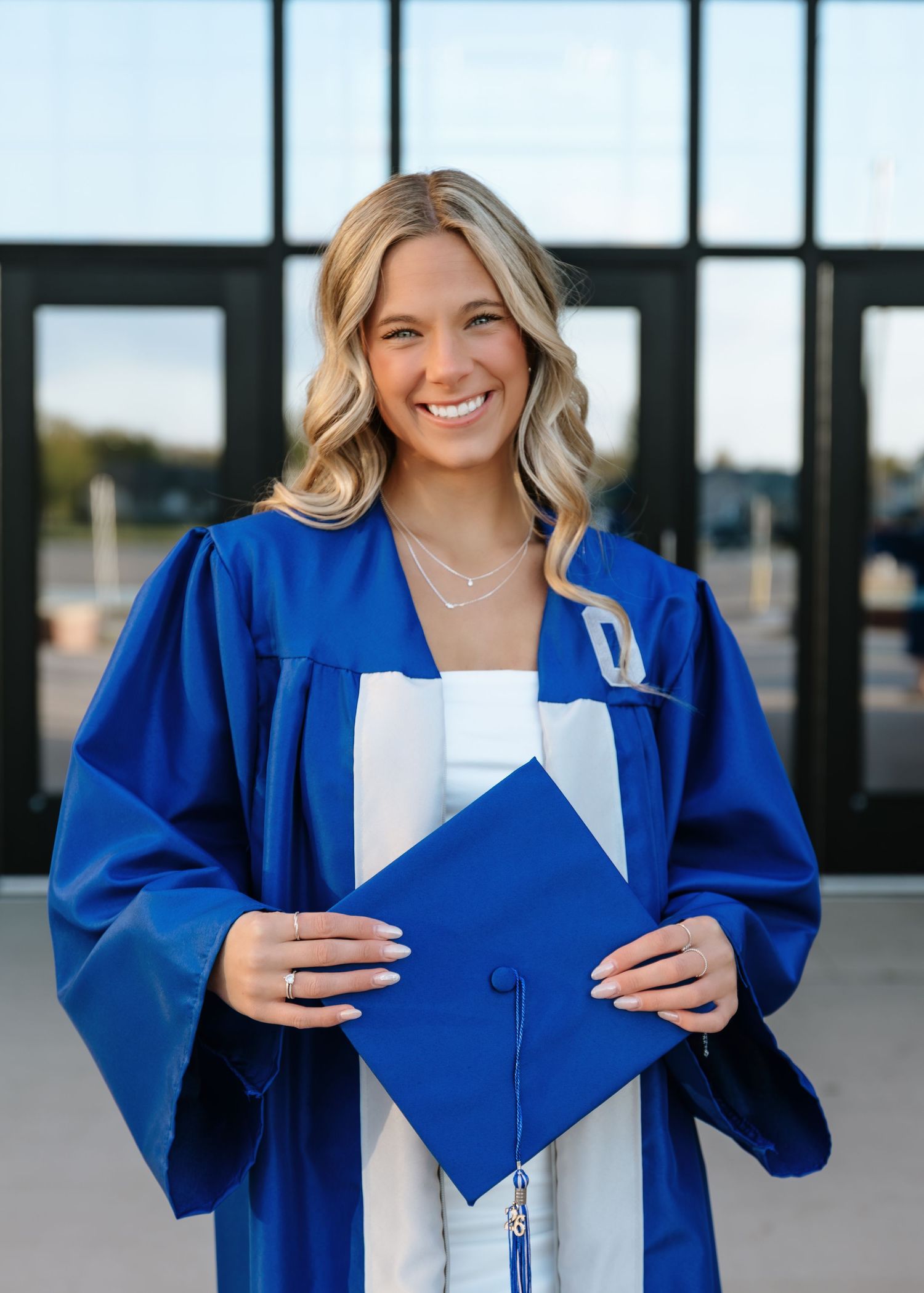 Smiling blonde female graduate in blue cap and gown holding mortarboard.