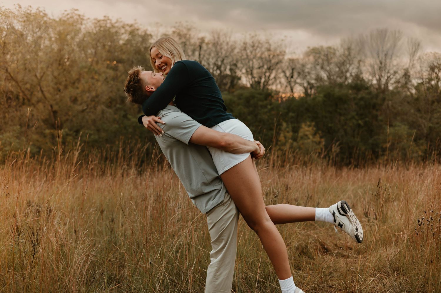 A romantic embrace in a golden field at sunset with tall grass and bare trees in the background.