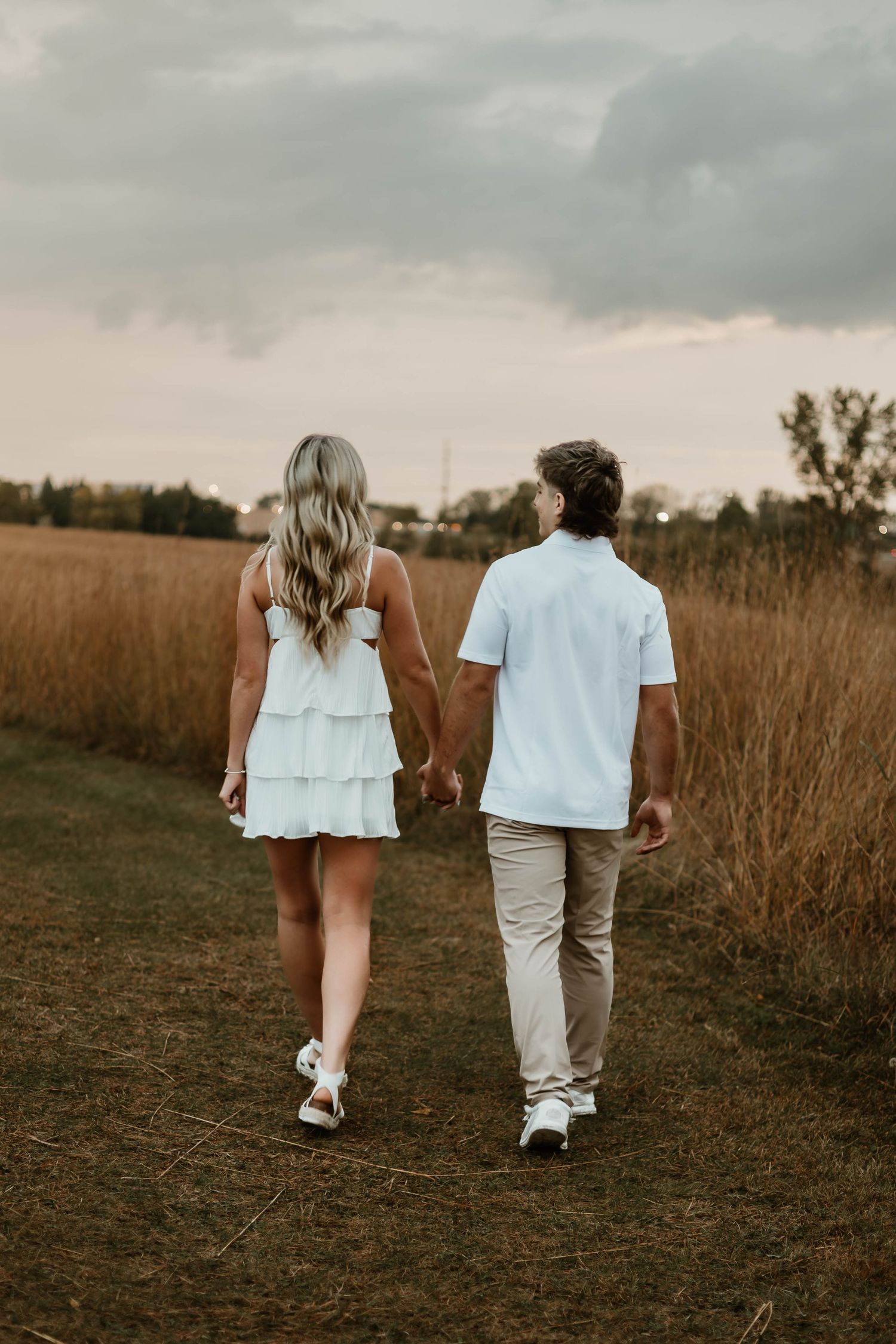 Couple walking hand in hand through a golden field at sunset, photographed from behind in matching white outfits.