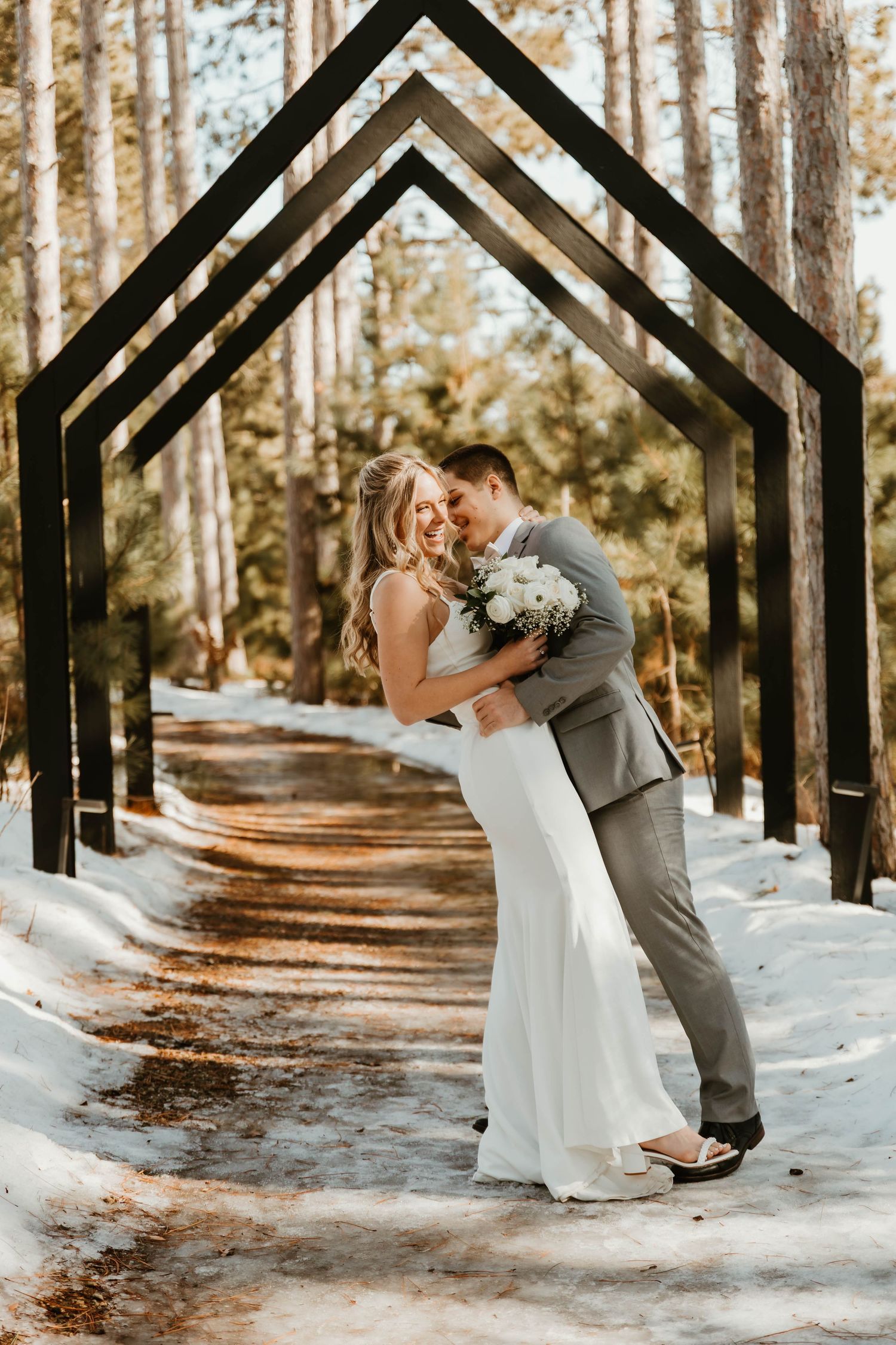 A romantic wedding moment under geometric arches on a snowy forest path.