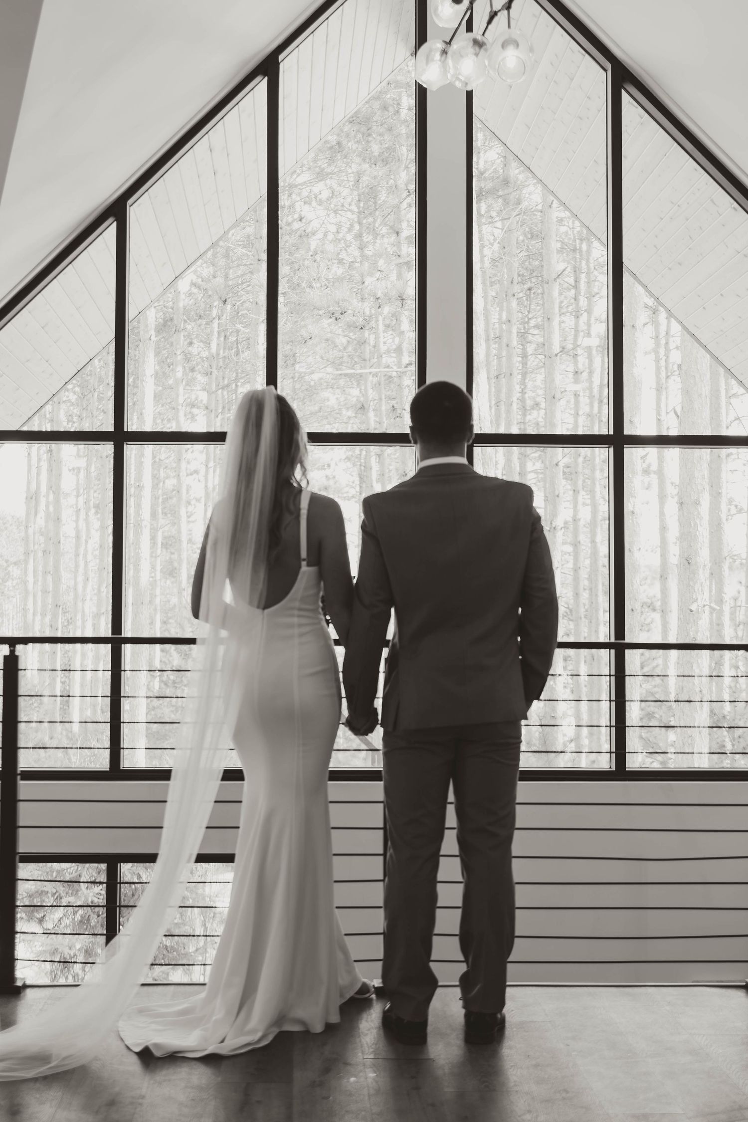 Silhouettes of a couple in wedding attire standing against large geometric windows with forest views.