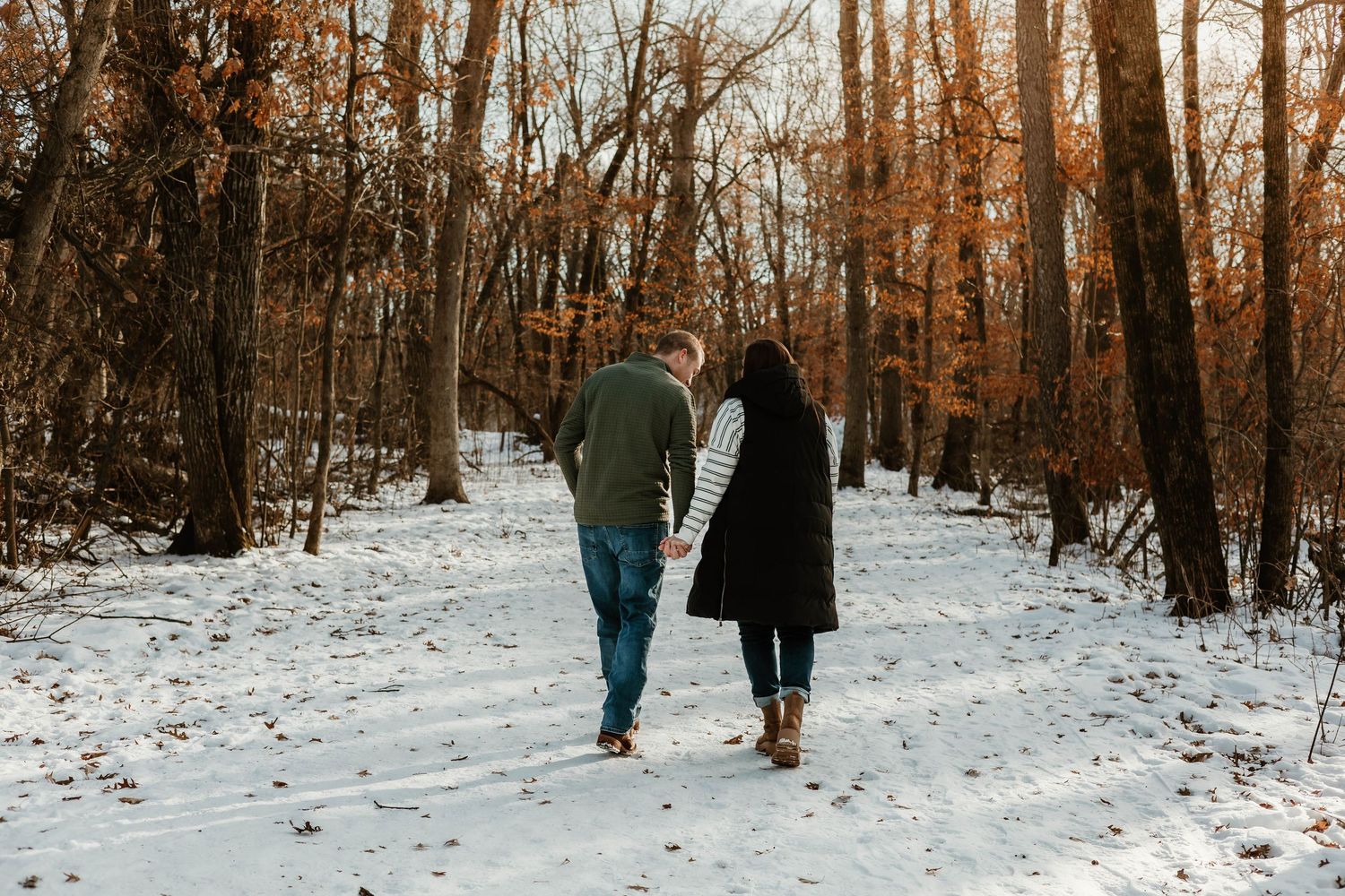 Two people walk through a snowy forest path surrounded by bare trees on a winter afternoon.