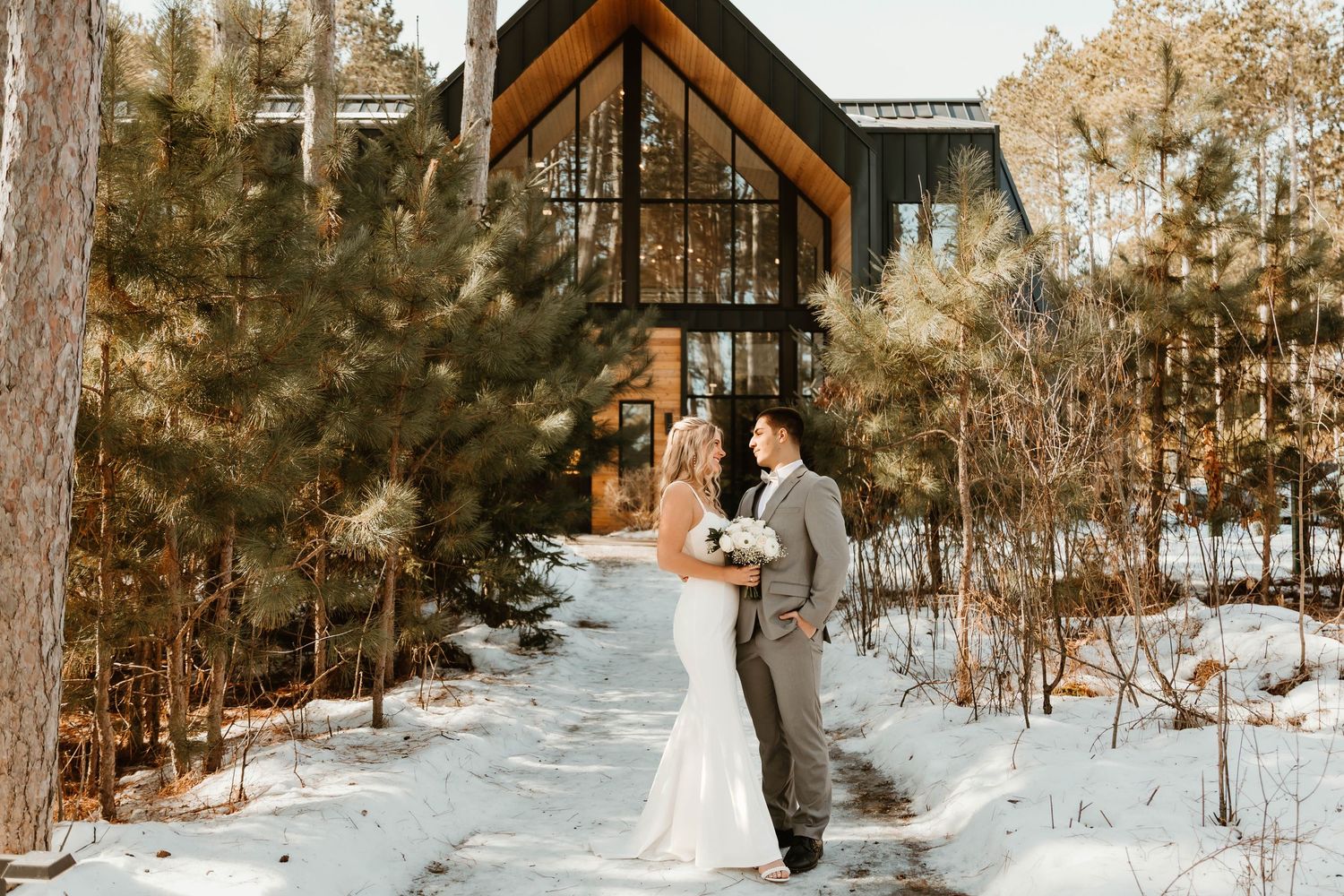 A couple stands together in front of a modern A-frame chapel surrounded by snowy pine trees in winter.