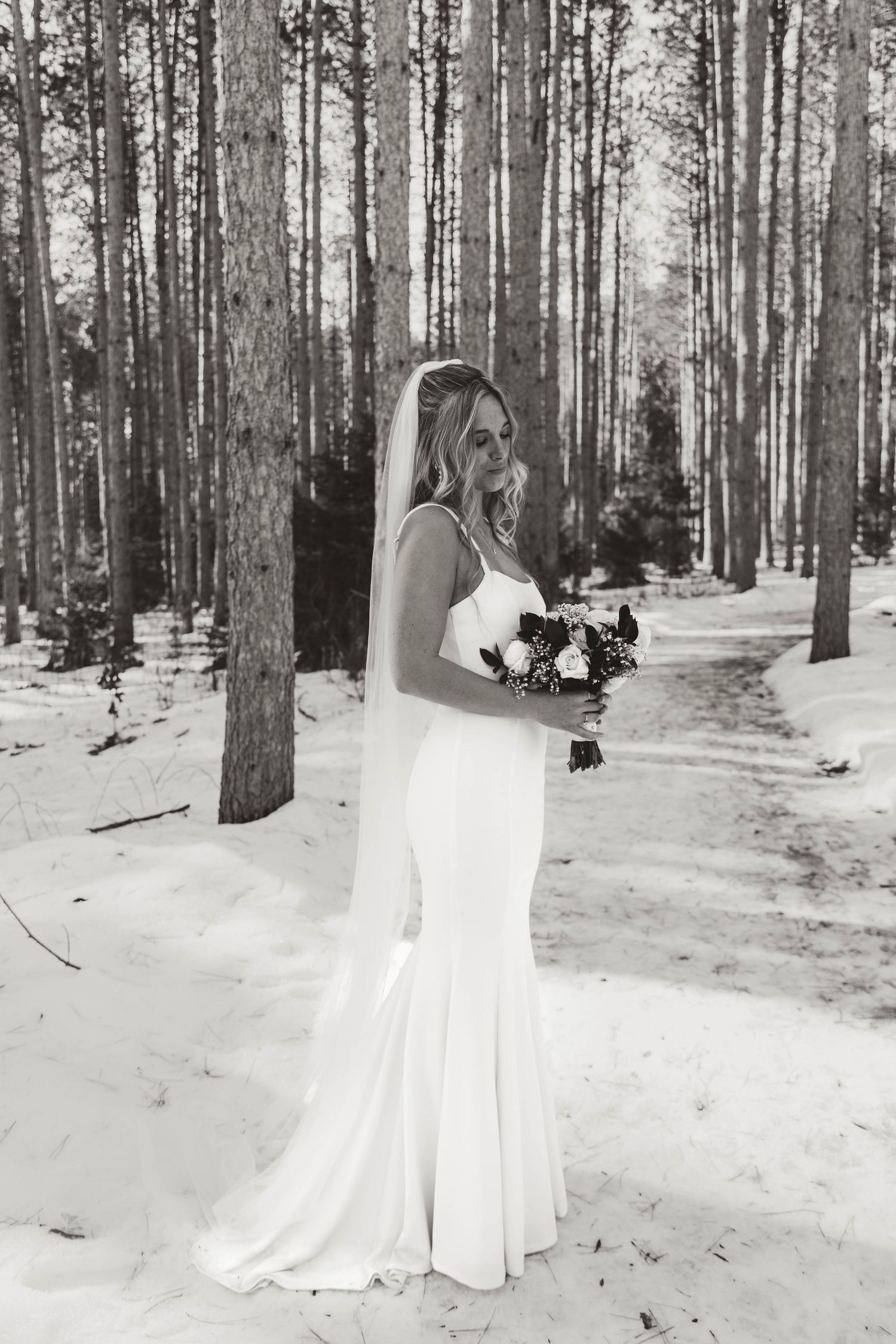 A bride in a white dress stands in a snowy forest holding a bouquet, captured in black and white photography.