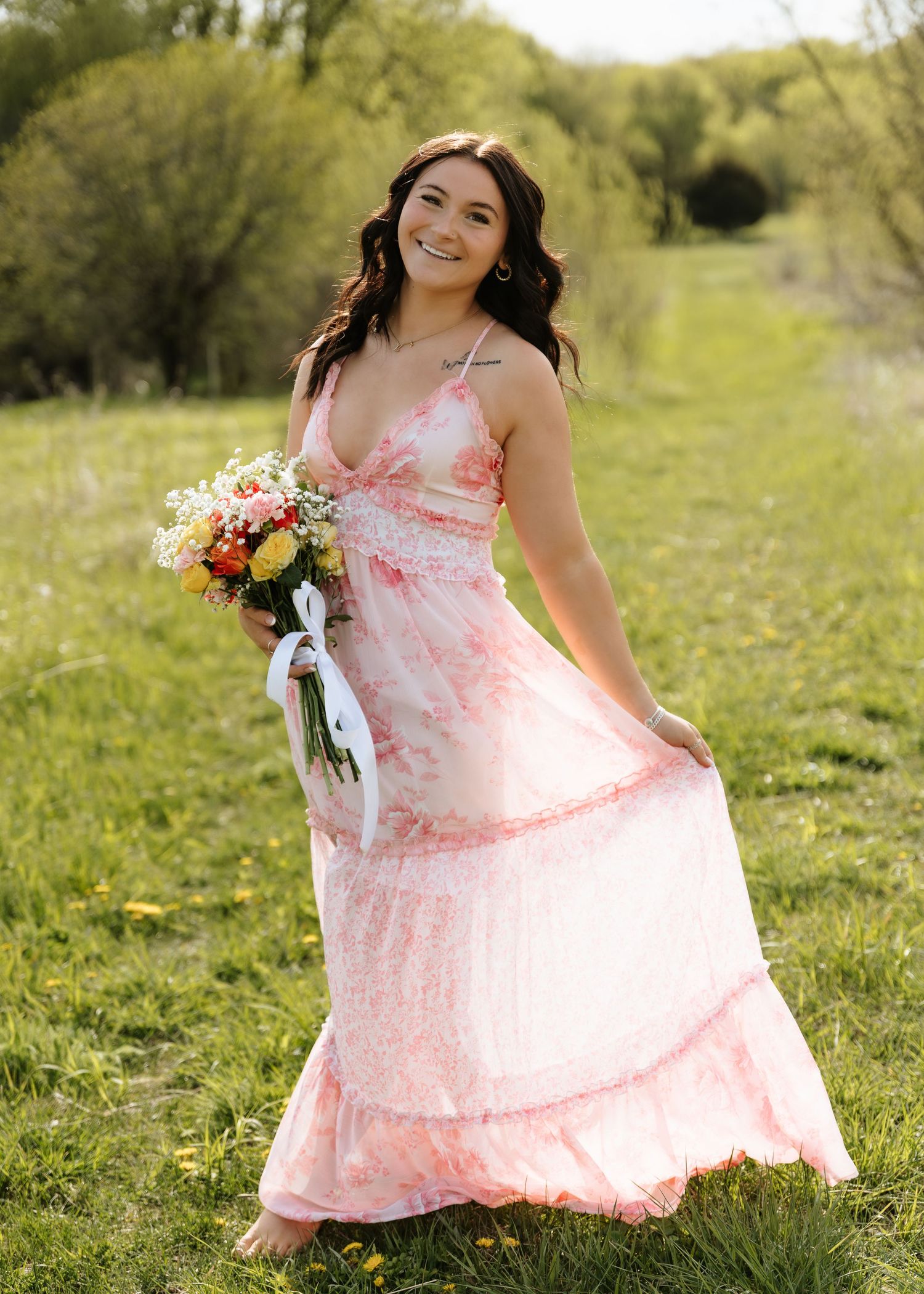 Young woman in pink floral maxi dress holding colorful bouquet, smiling in green meadow on sunny day.