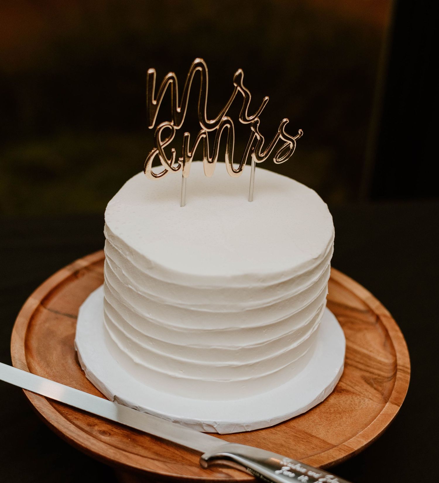 Simple white wedding cake displayed on wooden cake stand with metallic topper.