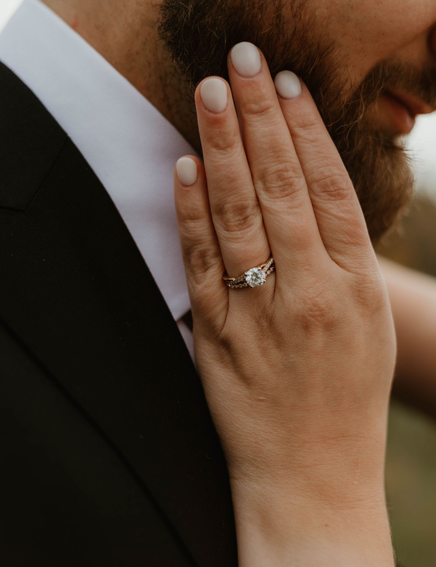 Close-up of a hand wearing an elegant diamond engagement ring against a dark suit jacket.