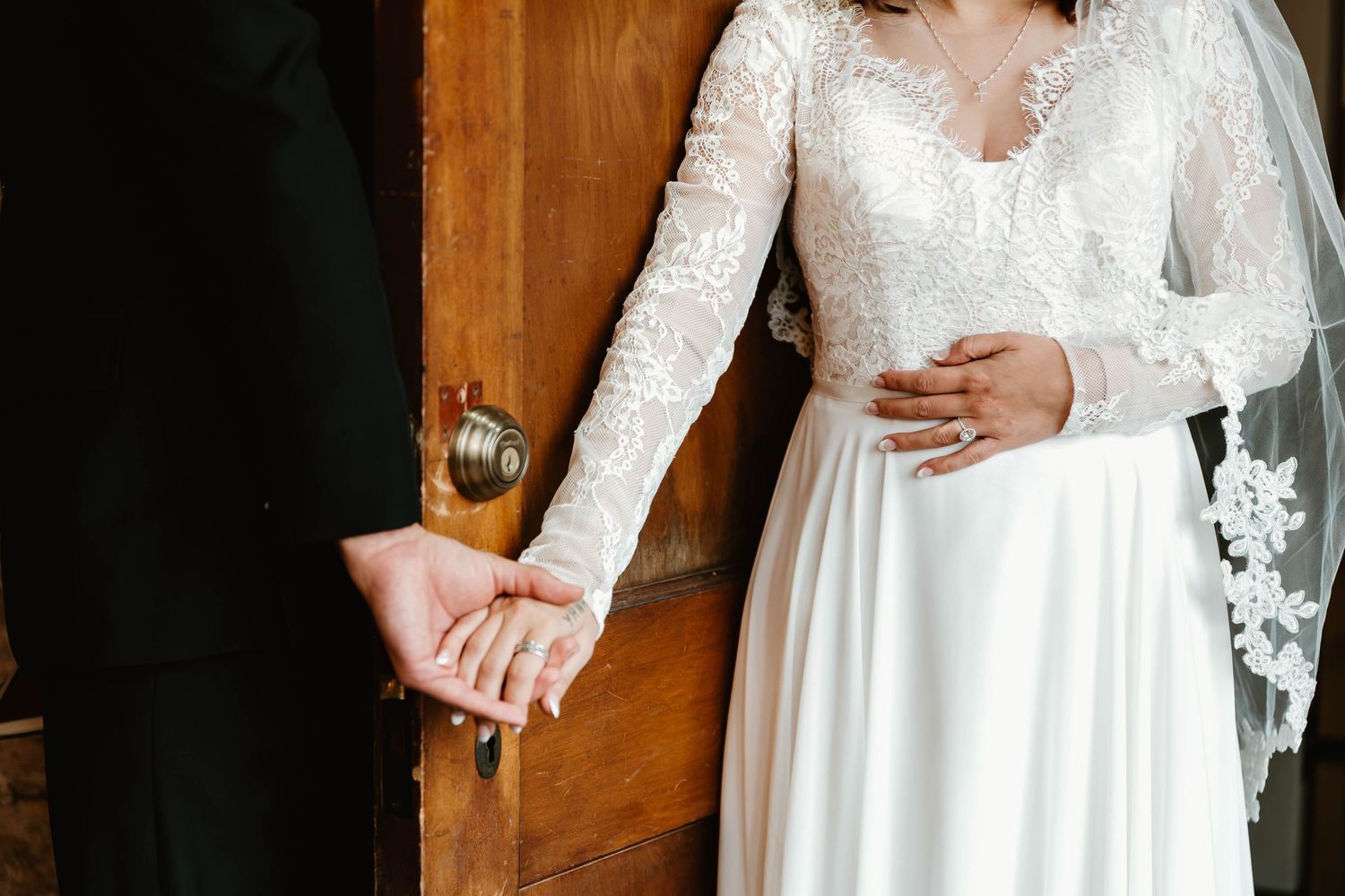 A bride in an elegant long-sleeved lace wedding gown holds a delicate white bouquet against a rustic wooden backdrop.