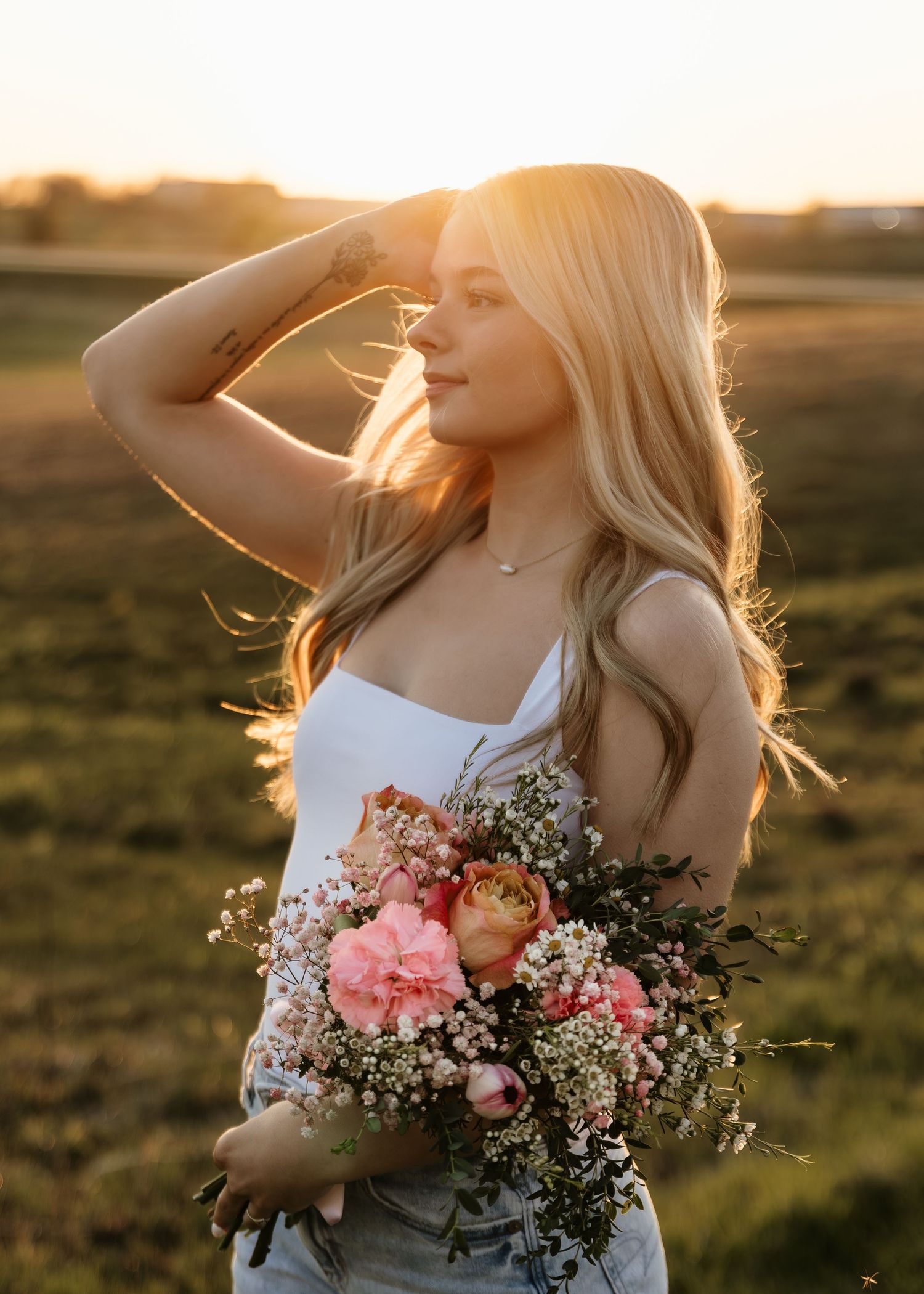 Young blonde woman holding a bouquet of pink flowers in a golden sunset field, wearing a white top.