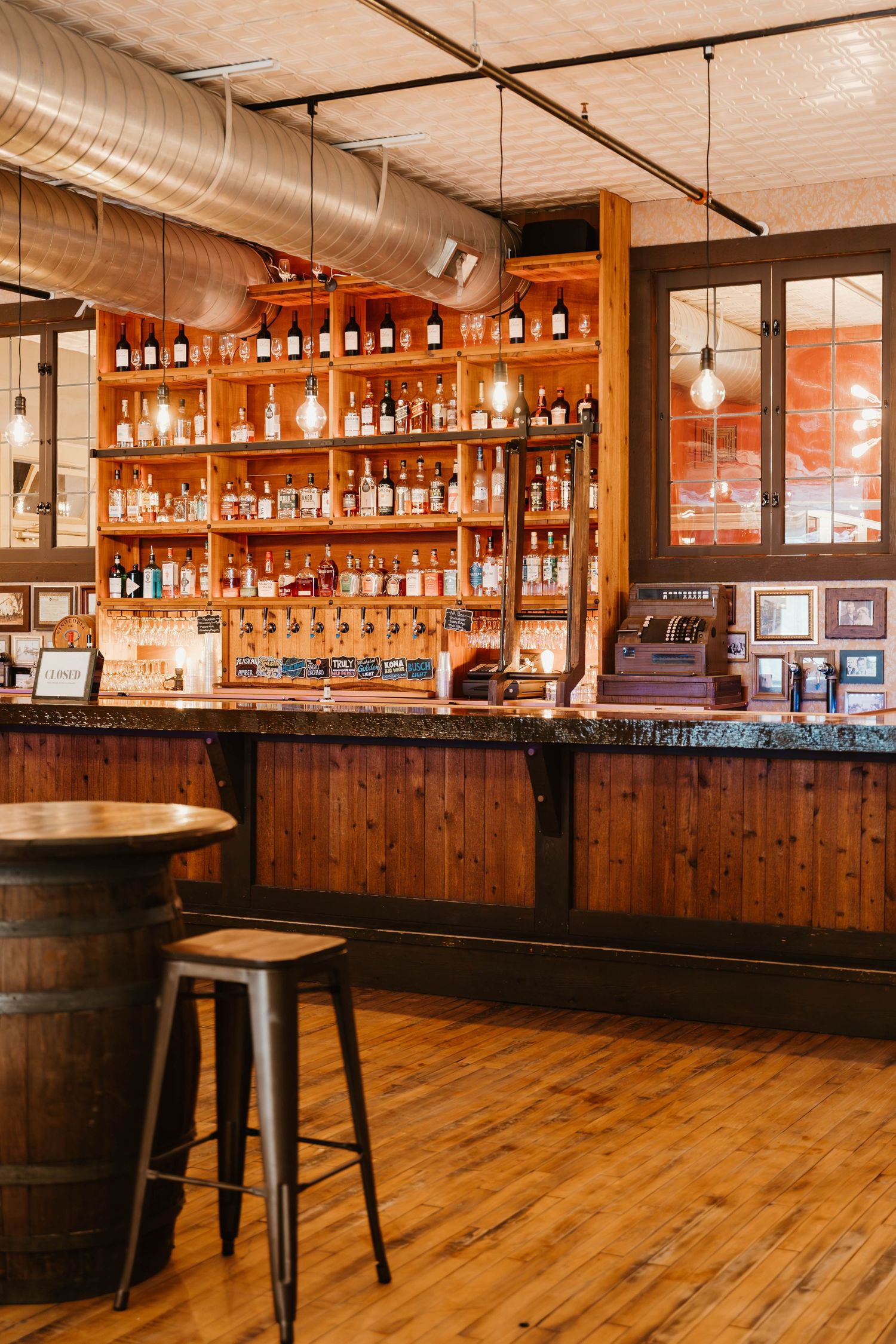 Rustic bar interior with wooden counters, exposed ductwork, and shelves lined with bottles.