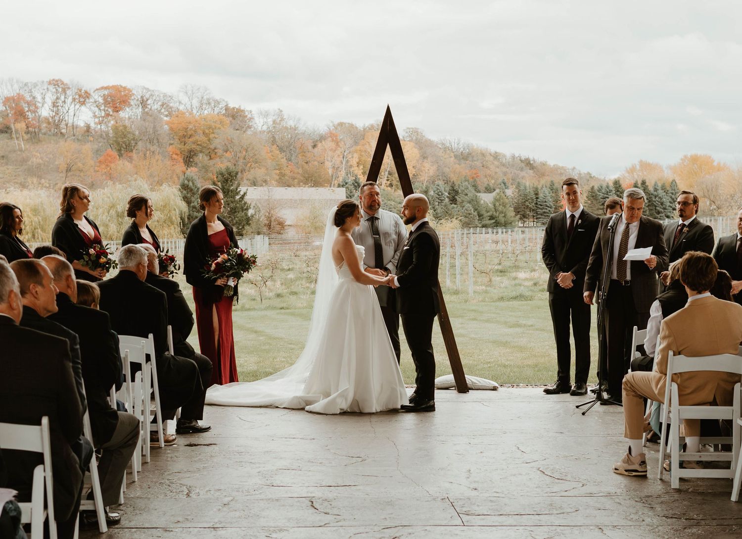 Outdoor autumn wedding ceremony with guests seated on white chairs overlooking fall foliage.