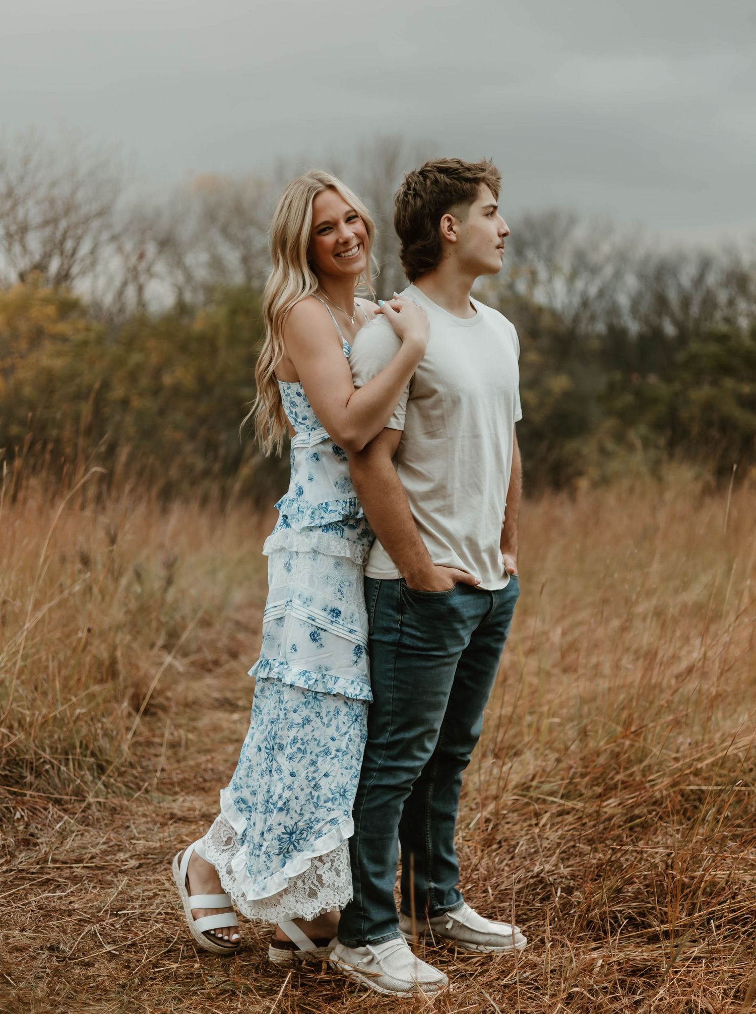 A romantic couple pose in coordinated outfits against tall grass and an overcast sky.