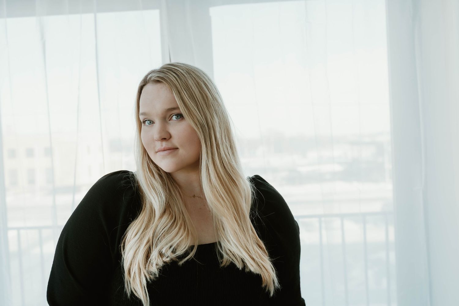 Professional portrait of a person in a black sweater against a bright window background.