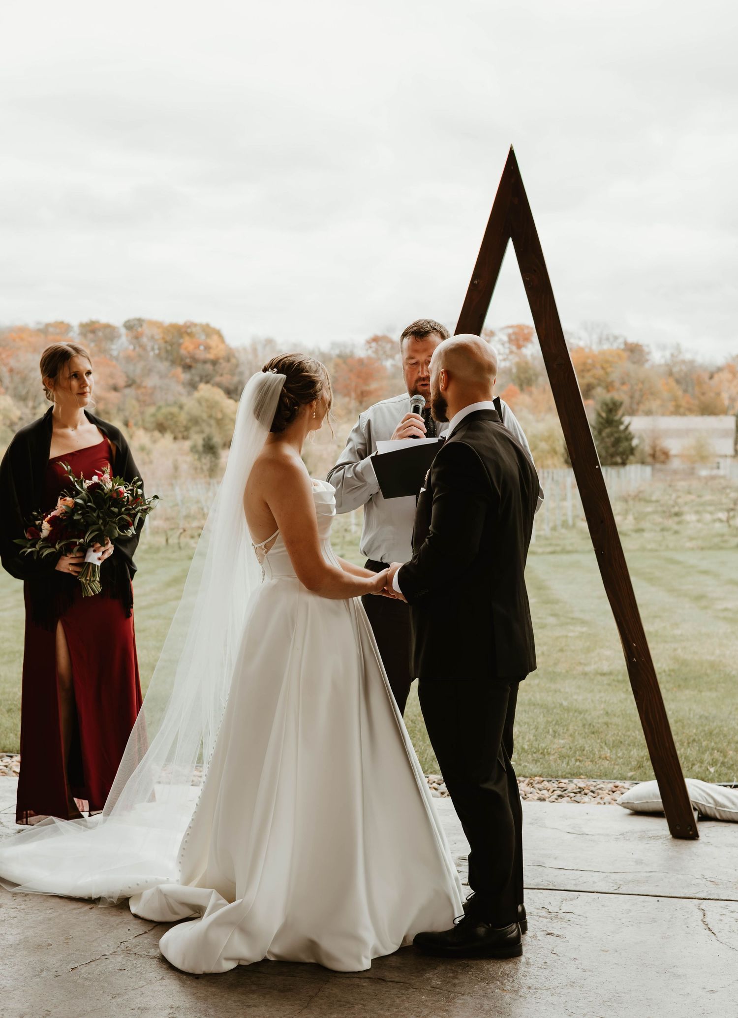 Outdoor wedding ceremony under wooden beams with fall foliage in the background.