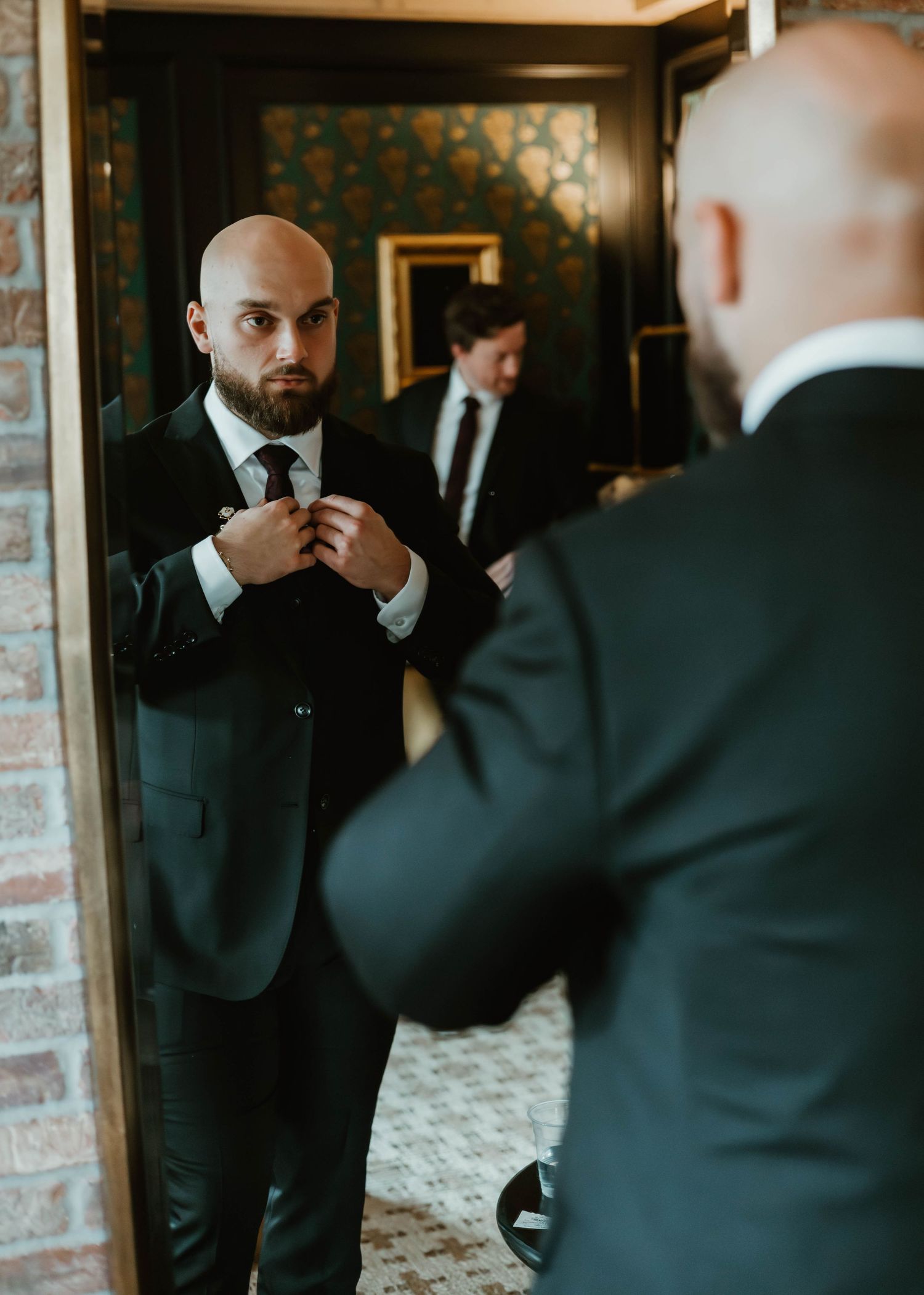 Groomsmen in black suits adjusting their ties and getting ready in front of a mirror before a wedding ceremony.