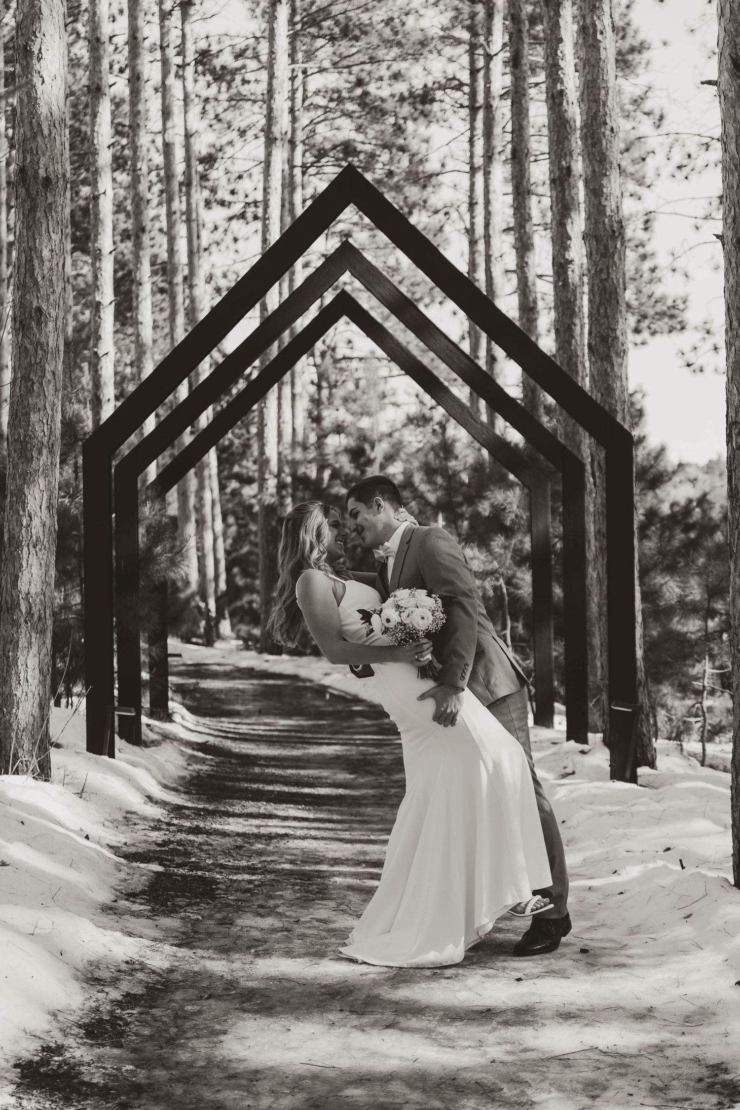 Black and white wedding photo showing a romantic dip under geometric arches in a snowy forest.