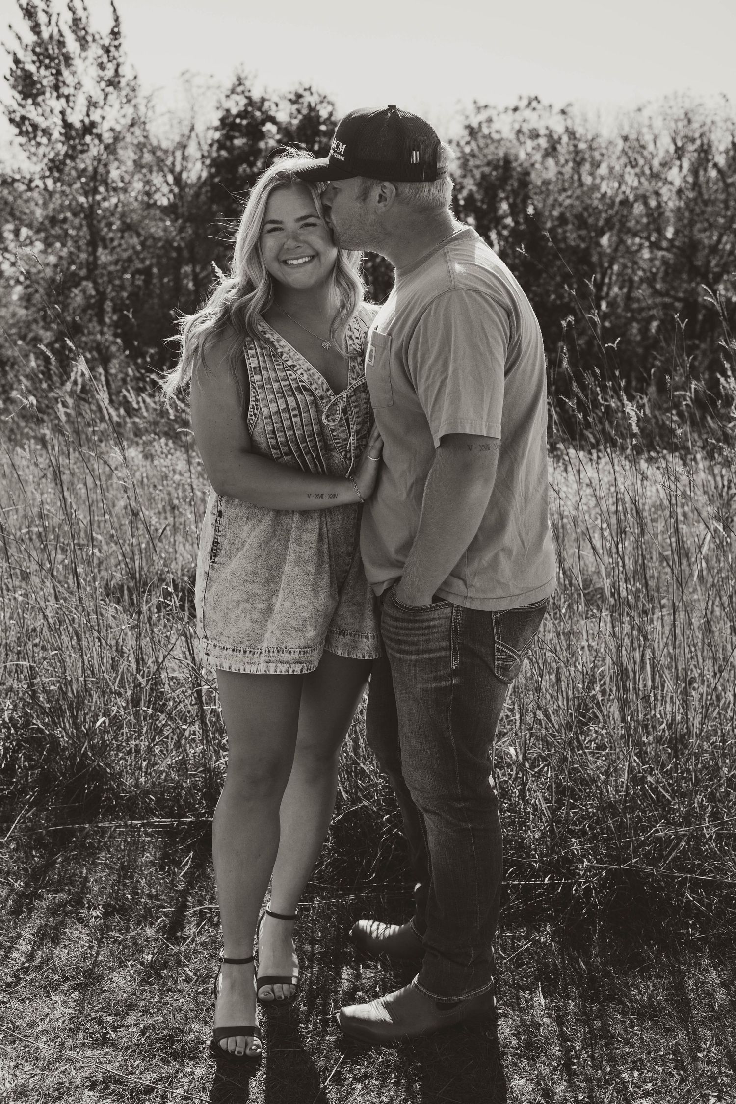 A couple shares an intimate moment together in a sunlit field with tall grass and trees in the background.