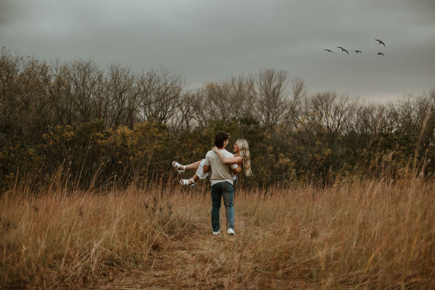 A couple walks through a golden autumn field at sunset holding hands while surrounded by tall dry grass.