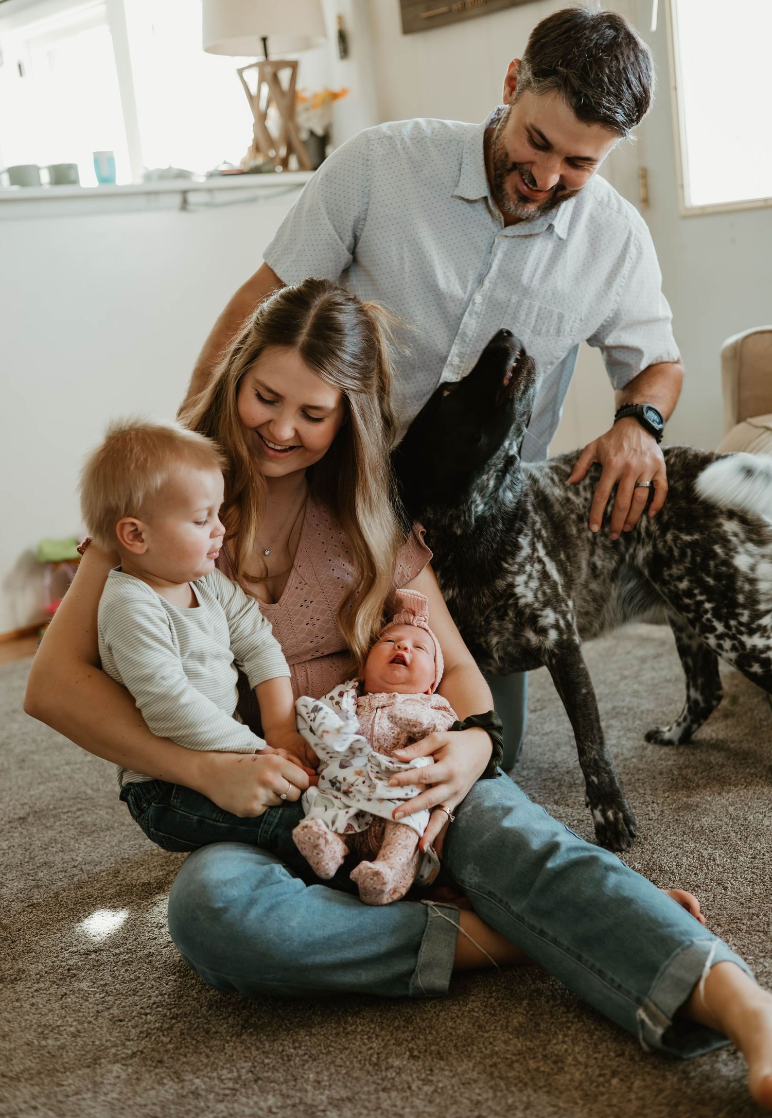Happy family with toddler, newborn baby, and spotted dog gathered together on living room floor.