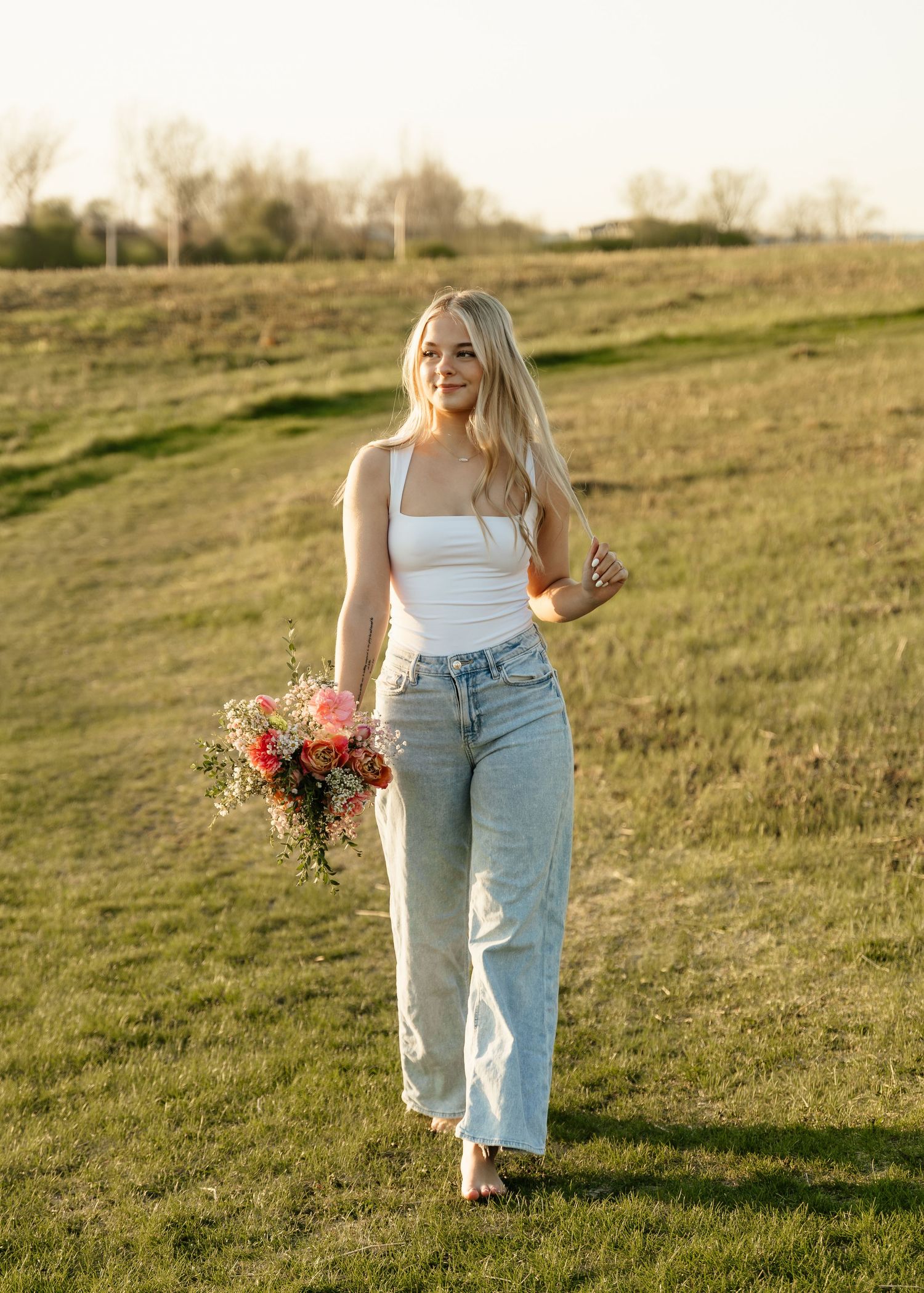 Smiling blonde woman in white top and light jeans holding colorful bouquet walking through green field at golden hour.