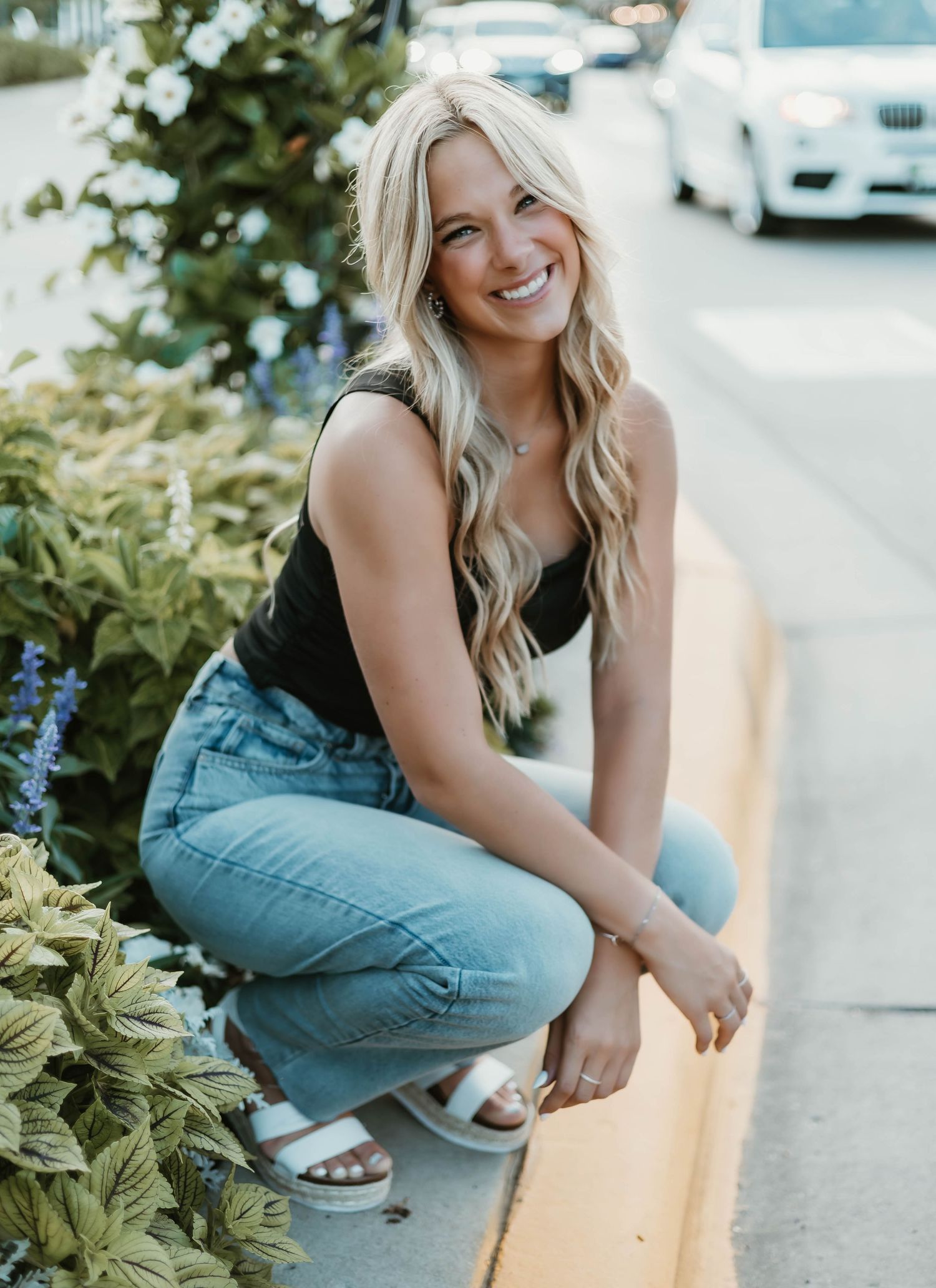 Smiling blonde woman crouching by colorful garden flowers on a sunny sidewalk, wearing jeans and a black tank top.