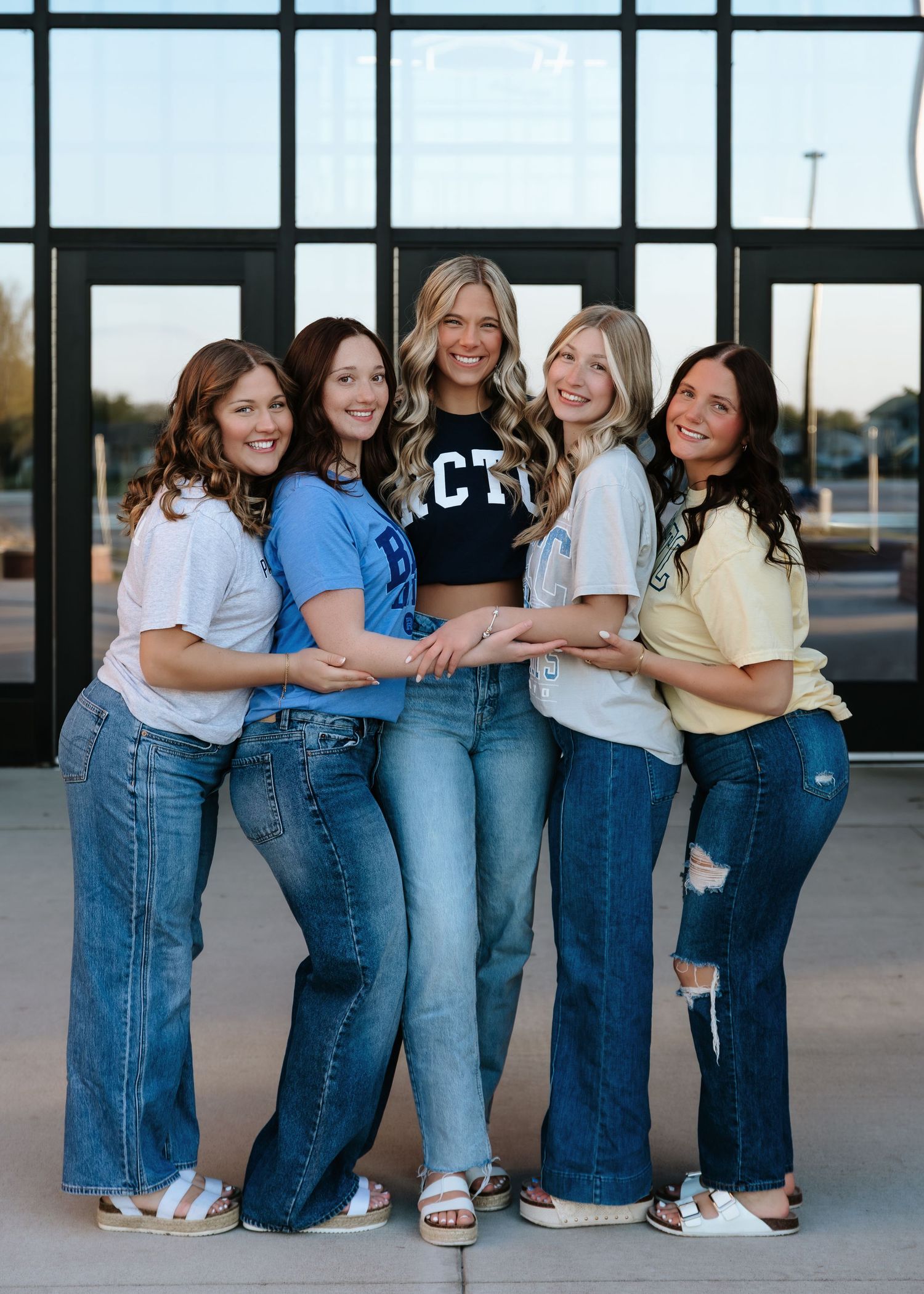 Five young women in jeans and casual tops smiling and posing together outside a modern building.