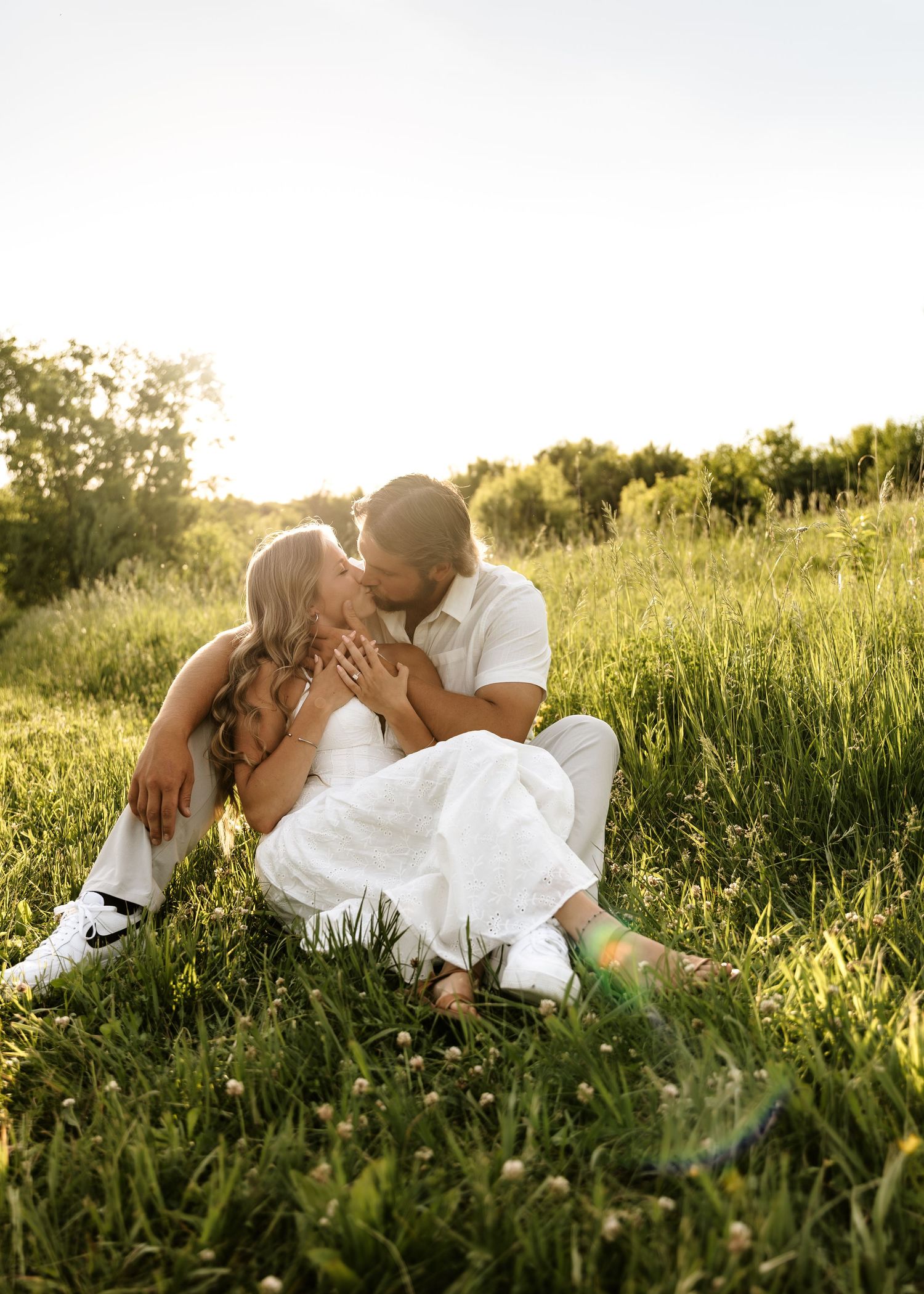 Couple sharing a romantic kiss while sitting in a lush green meadow during golden hour in wedding attire.