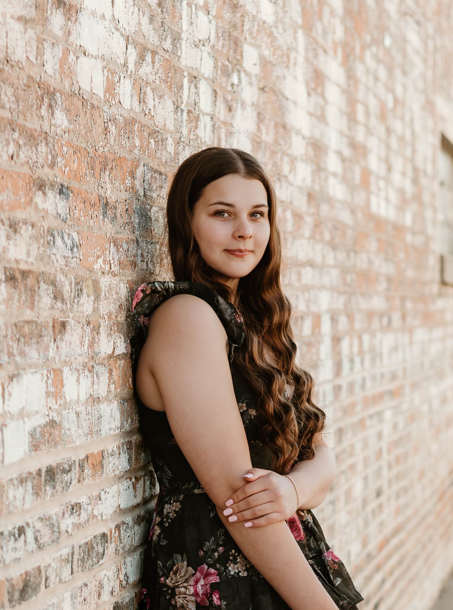 Young woman with long wavy brown hair in floral dress leaning against rustic brick wall.