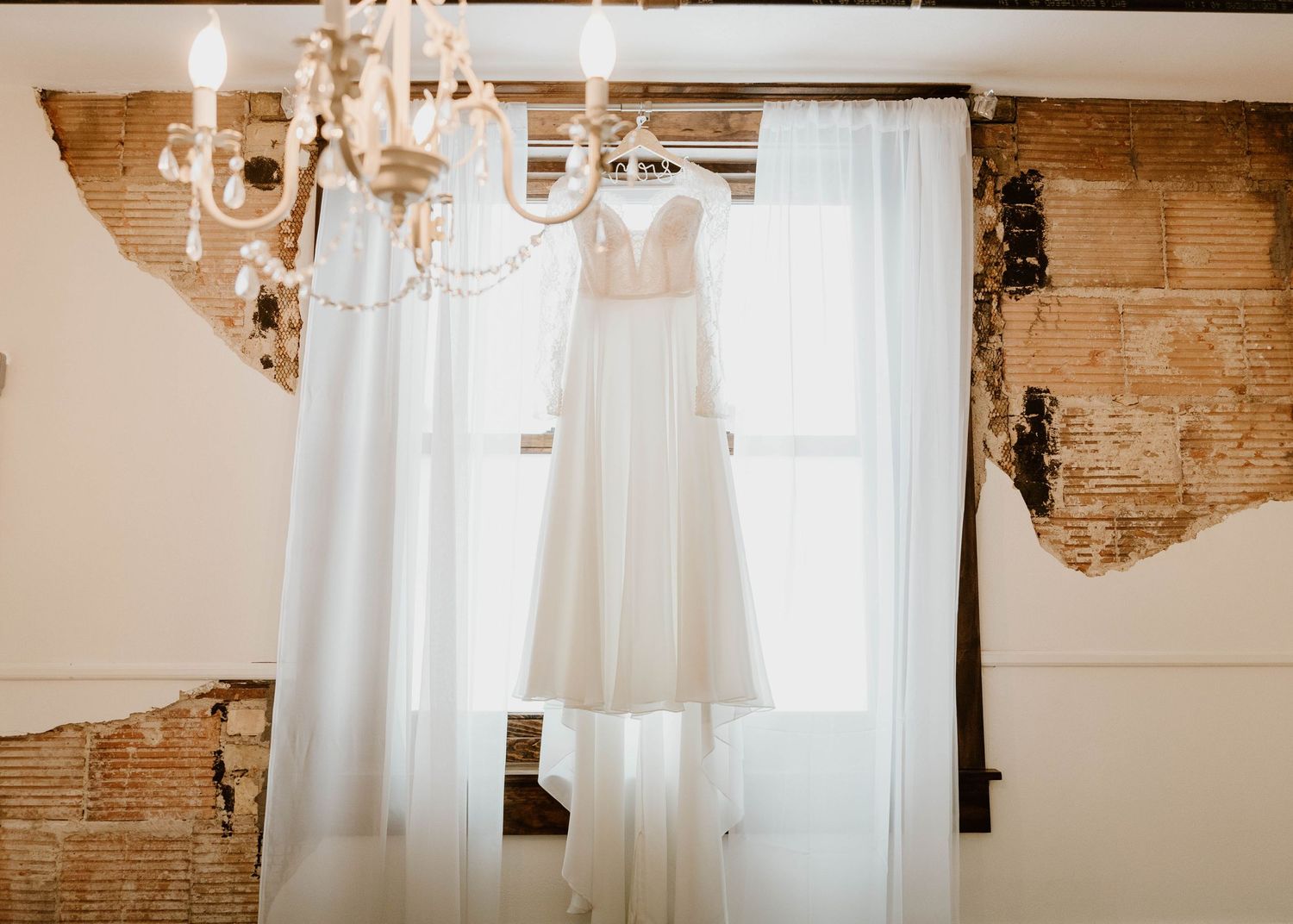 A white wedding dress hangs from an elegant chandelier in a rustic room with exposed brick walls and sheer curtains.