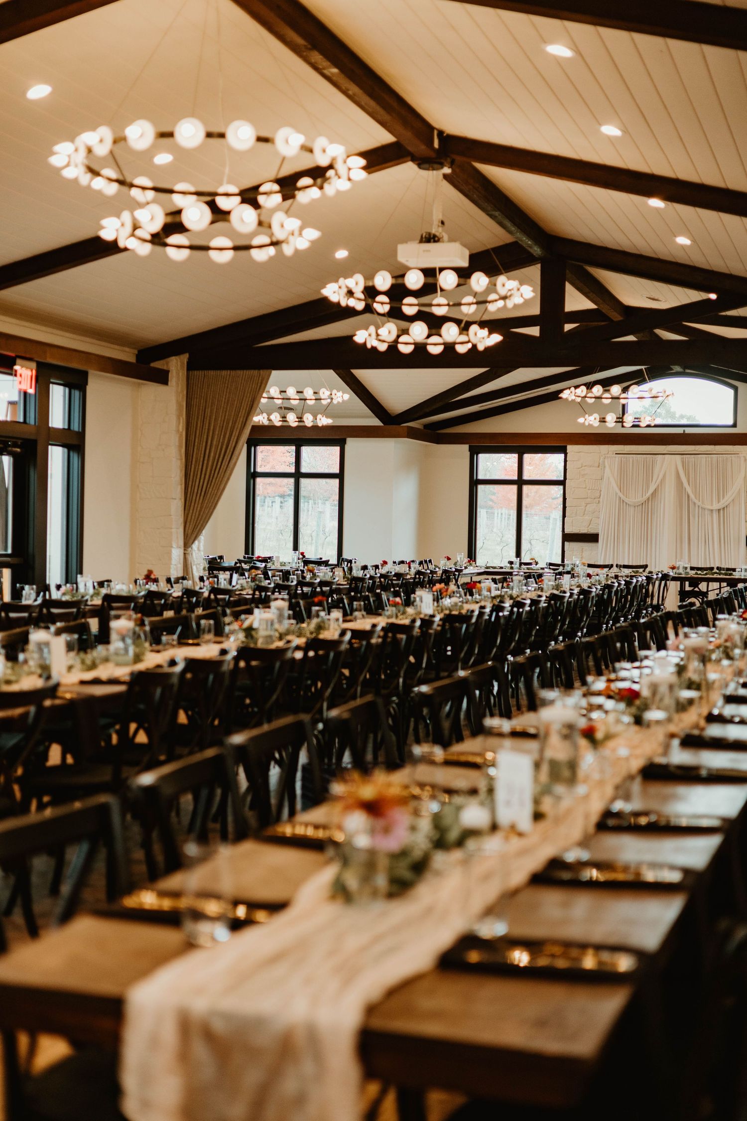 Elegant wedding reception venue with long tables and chandeliers under wooden beams.