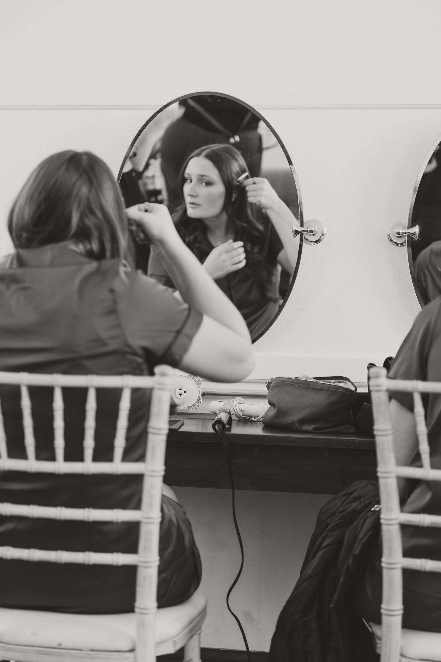 Black and white image of bridesmaids getting their hair styled while seated in white chairs.