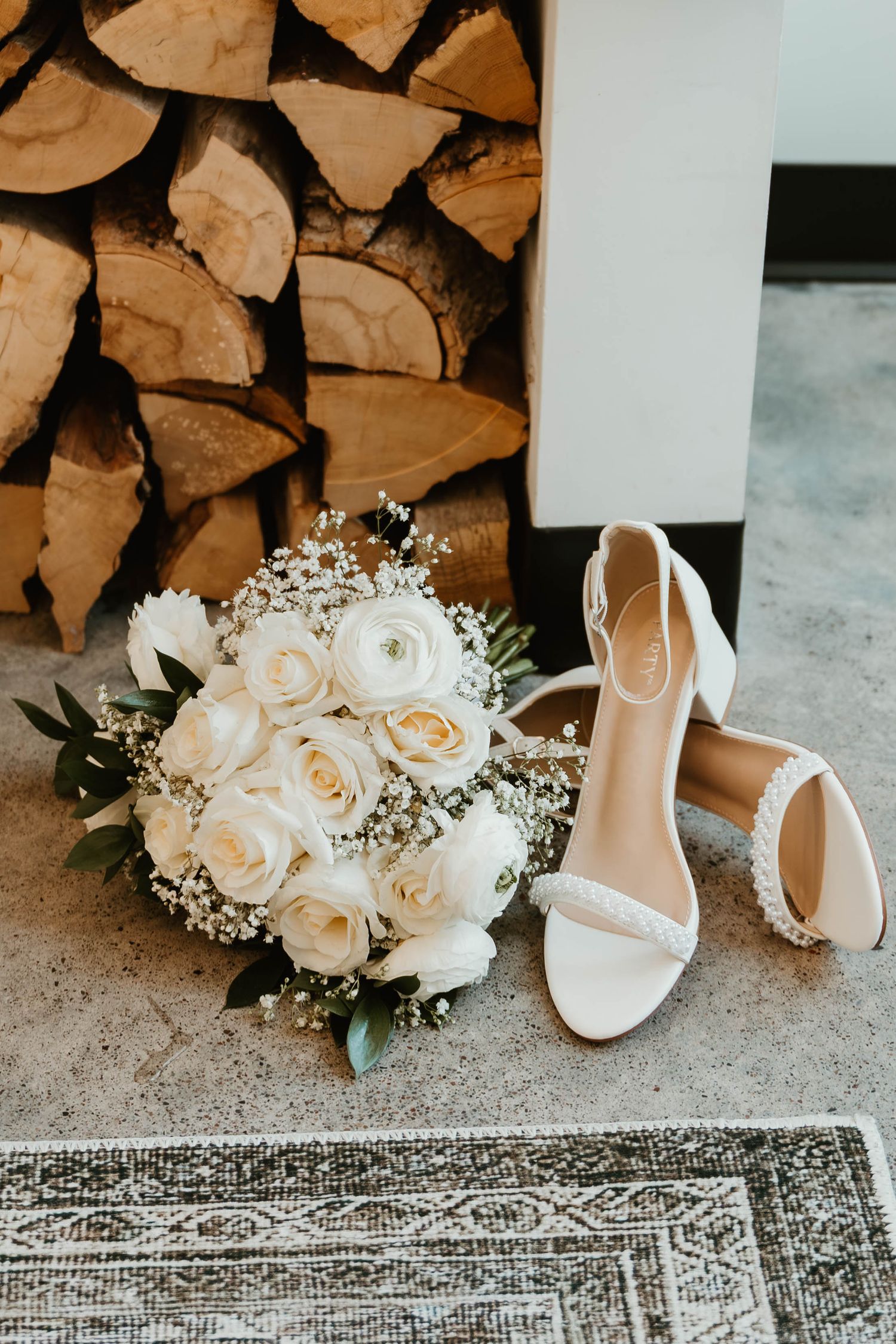 White bridal shoes and roses arranged beside stacked firewood and a vintage rug.