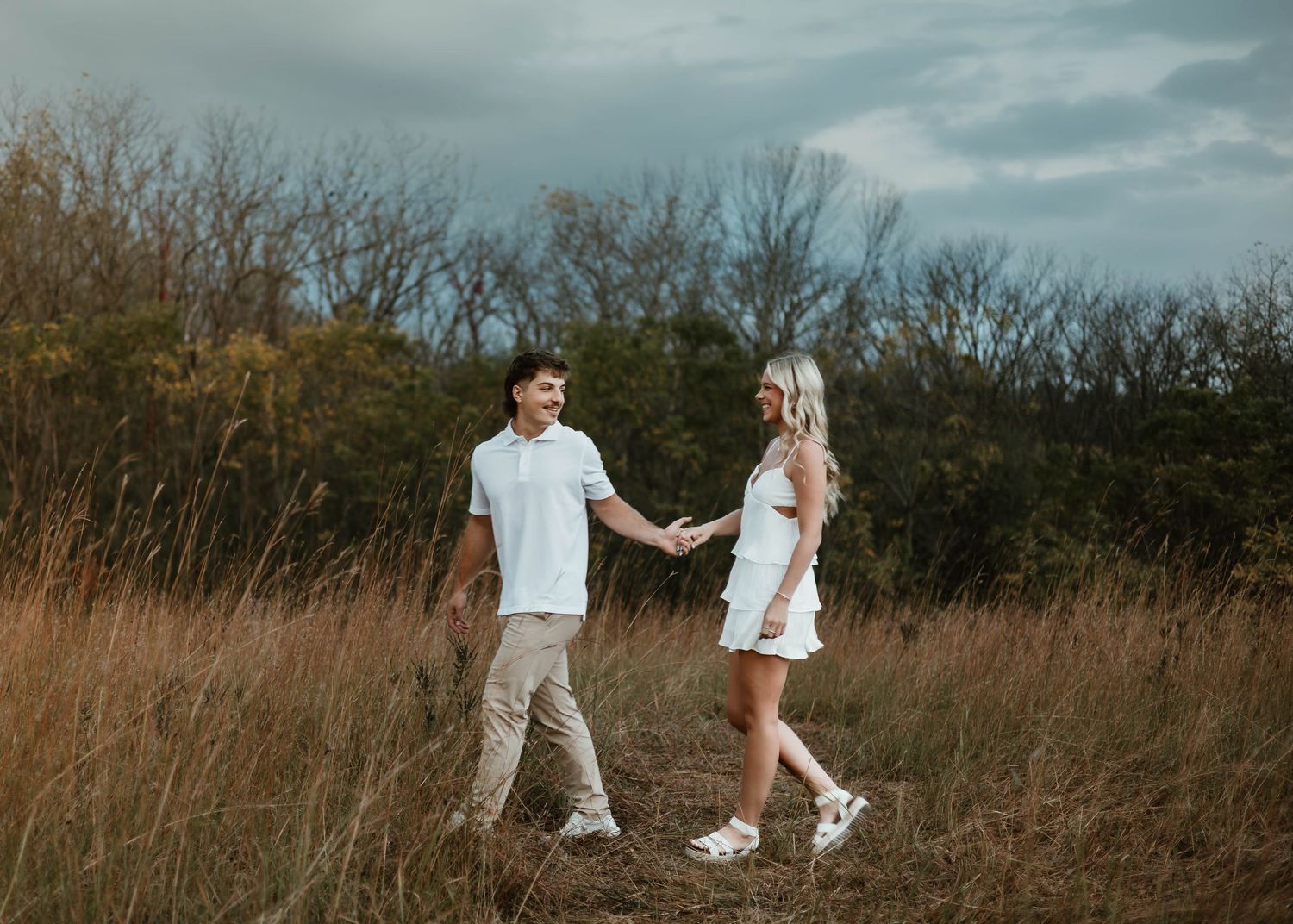 A couple in white outfits walks through a rustic field with tall grass at sunset.