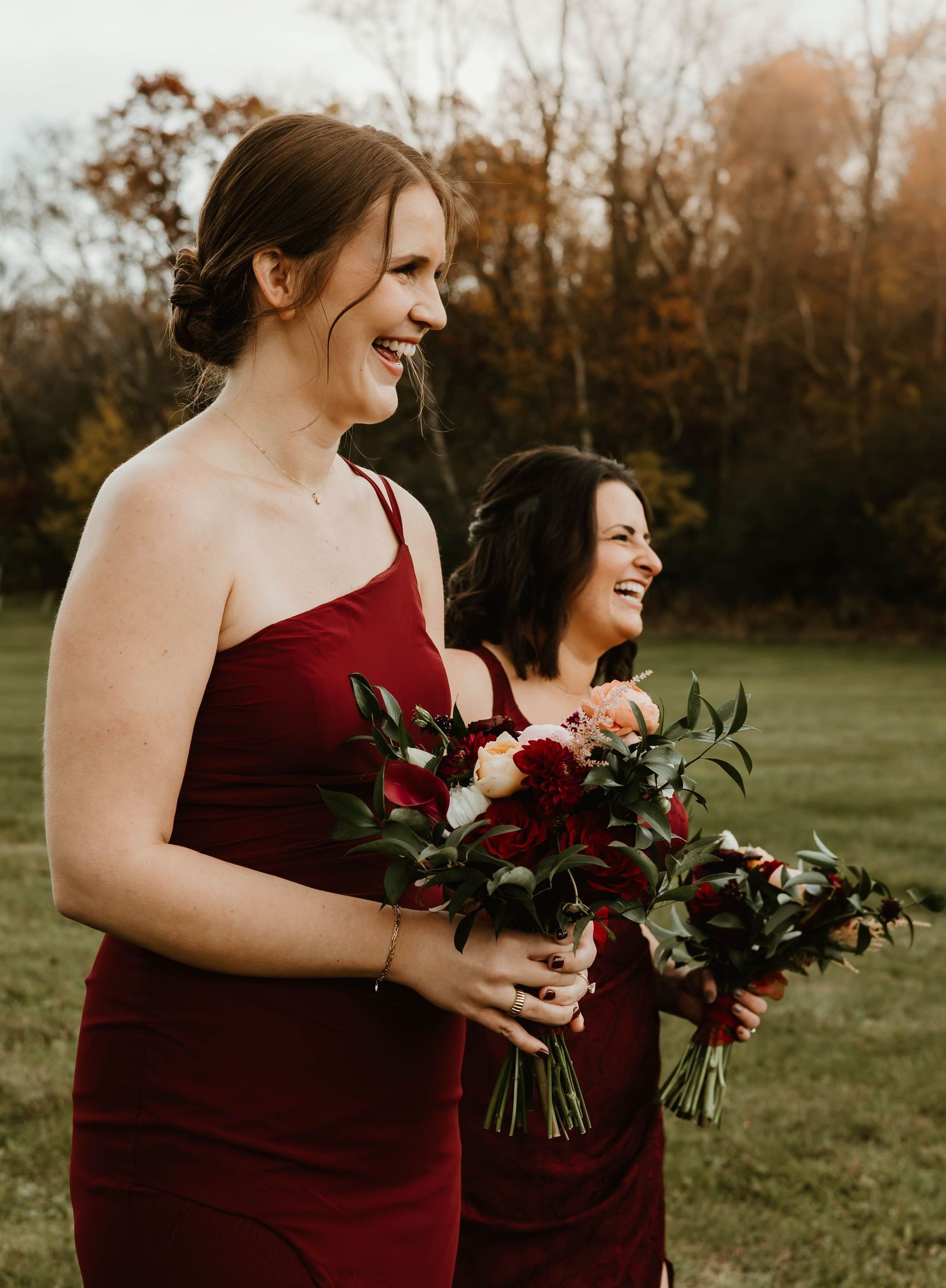 Bridesmaids in burgundy dresses hold bouquets of deep red flowers and greenery in an autumn setting.