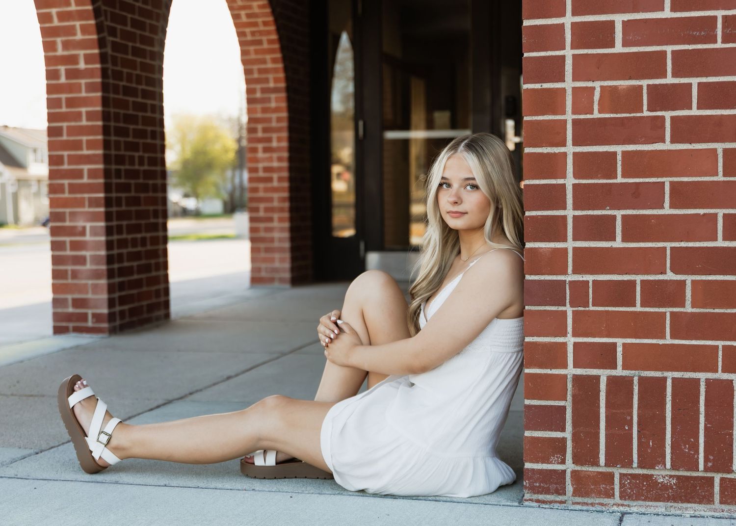 Young blonde woman in white dress and sandals sitting against brick wall outdoors for senior portrait photo.
