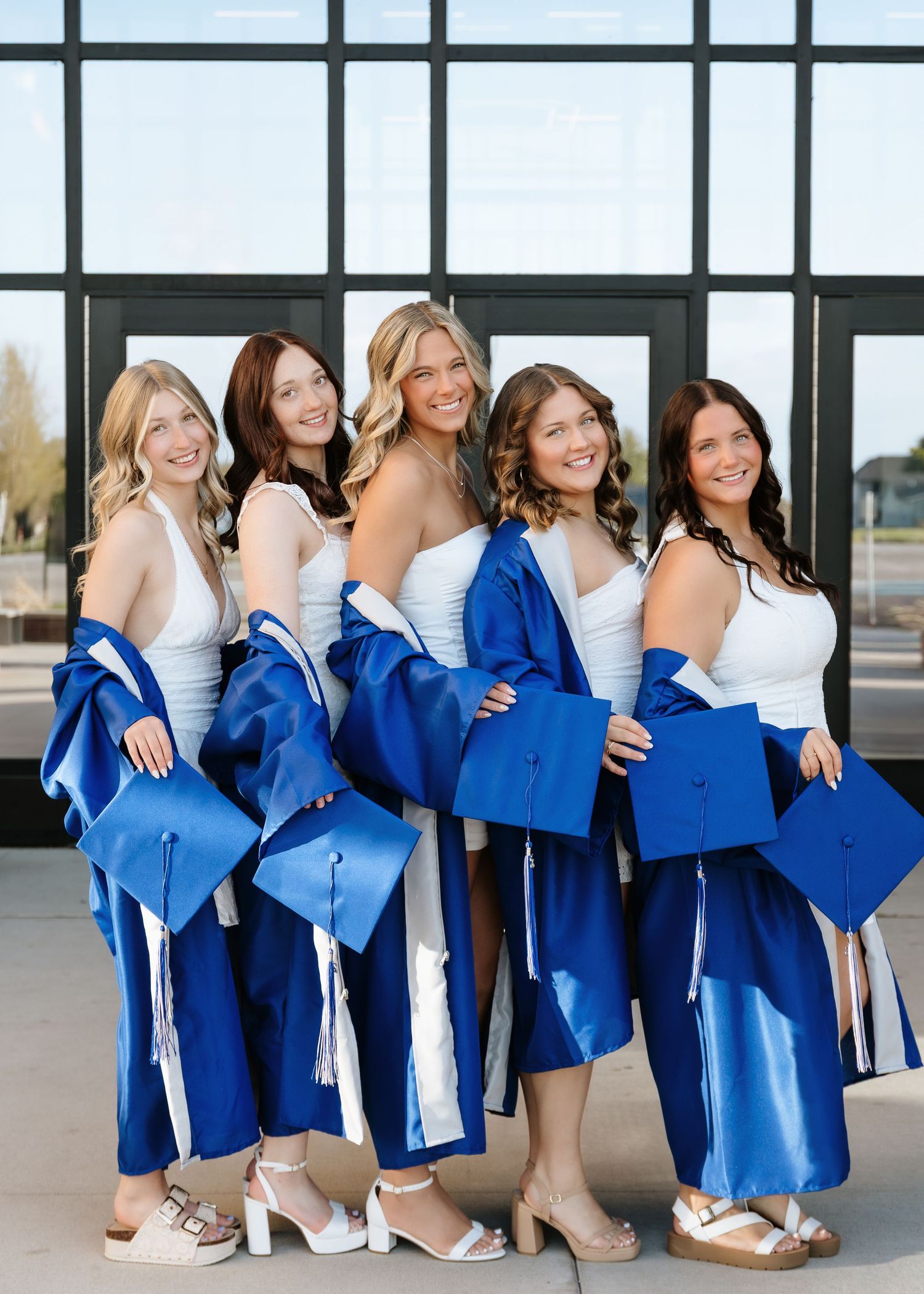 Five young women in white dresses pose with royal blue graduation caps and gowns, celebrating their graduation day.