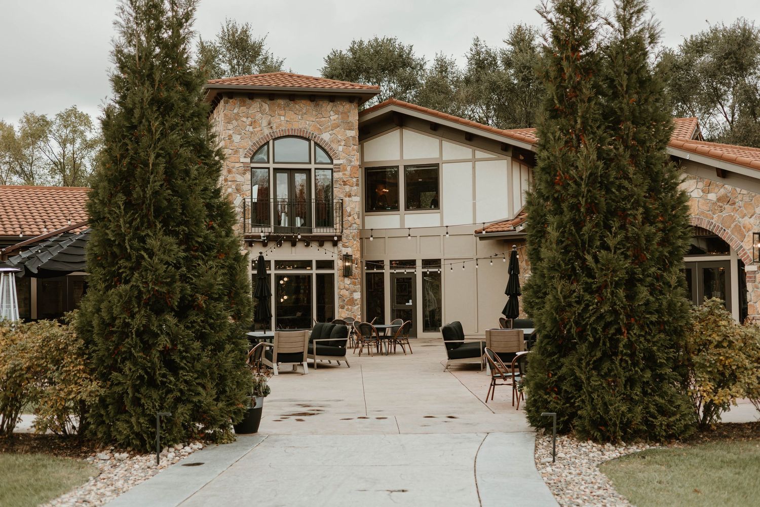 Modern stone and wood home exterior with tall windows and cypress trees lining a concrete walkway.