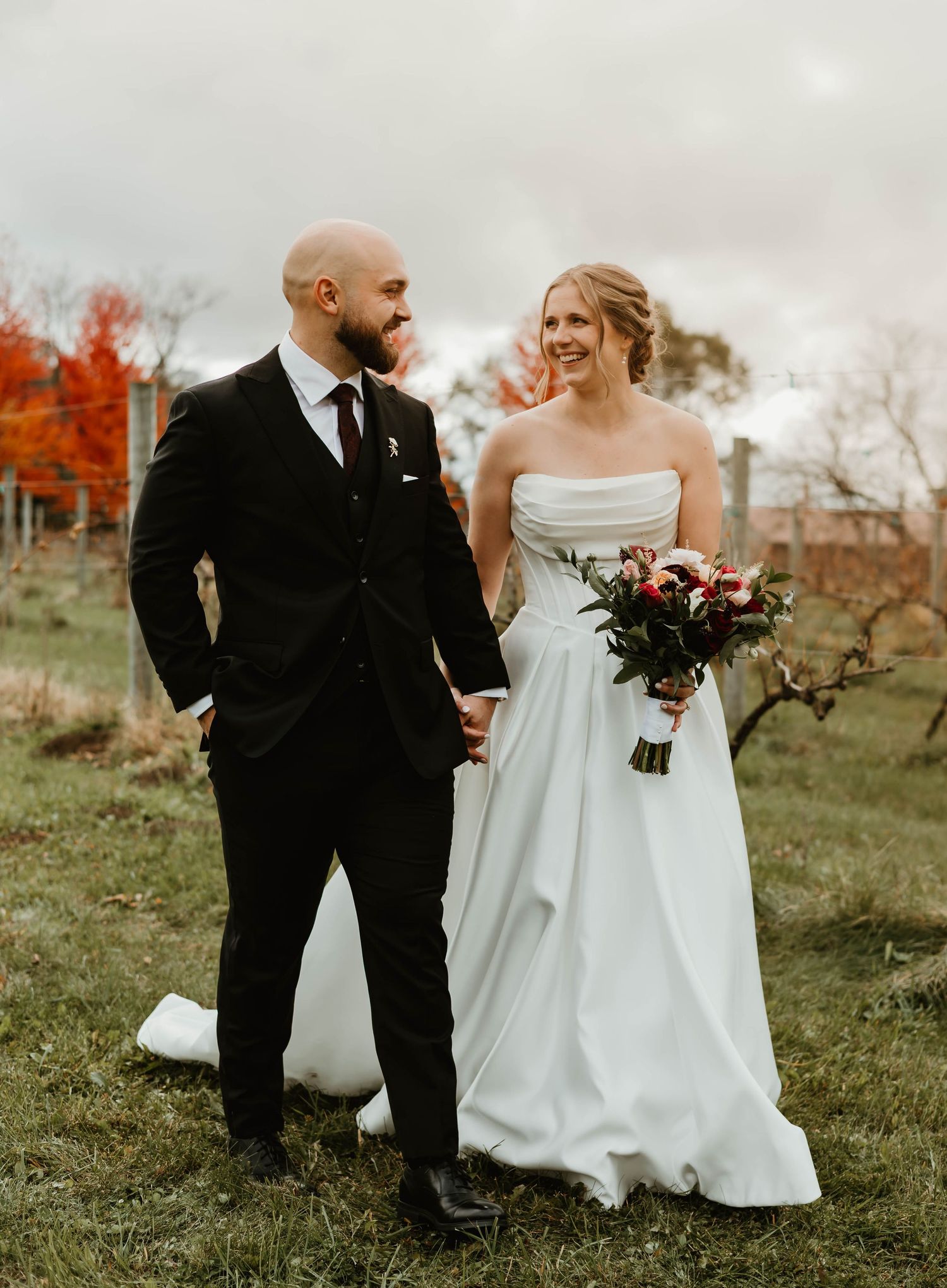 Couple in wedding attire walking through autumn field with rustic fencing.