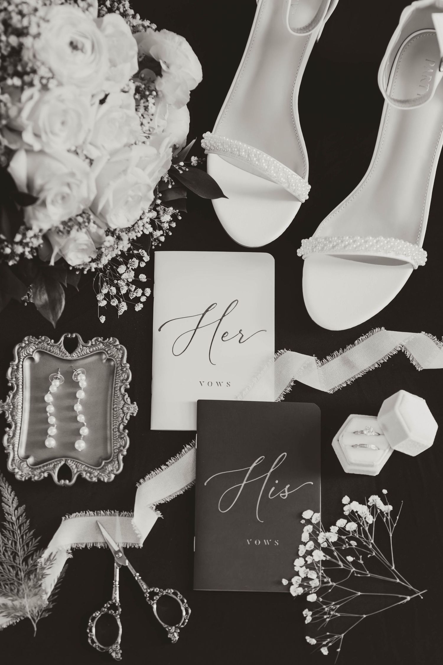 Wedding details flatlay with white shoes, vow books, roses, and pearl accessories on black background.