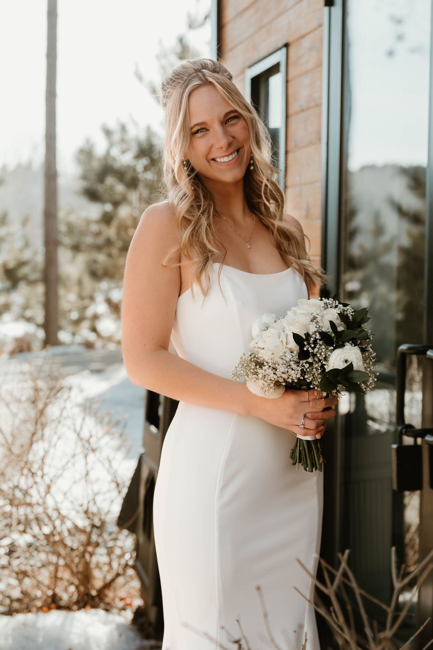 A bride in a white dress holds a delicate bouquet of white flowers and greenery against a modern building exterior.