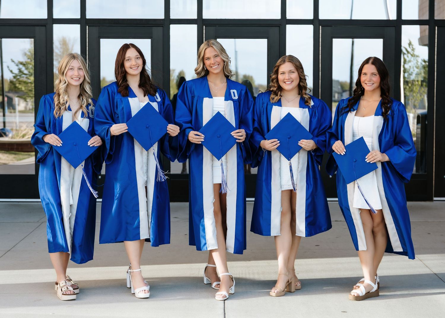 Five women in blue graduation gowns and caps smile together outside a modern building.