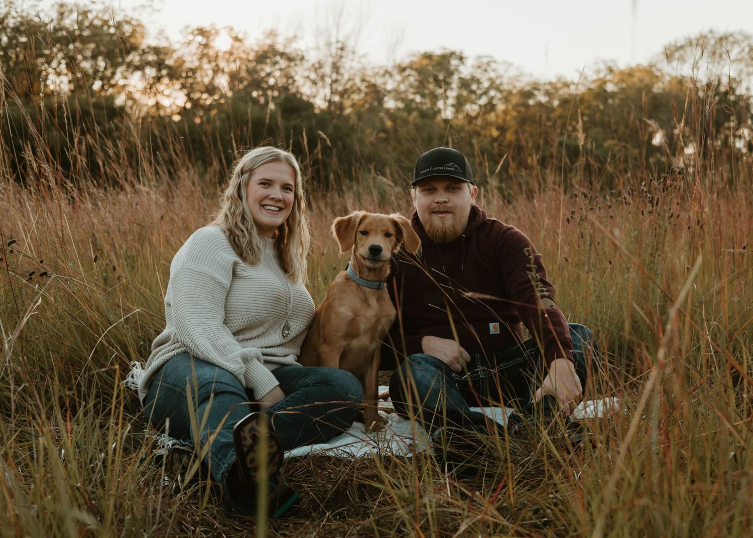 A couple and their dog share tender moments in a golden autumn field at sunset.