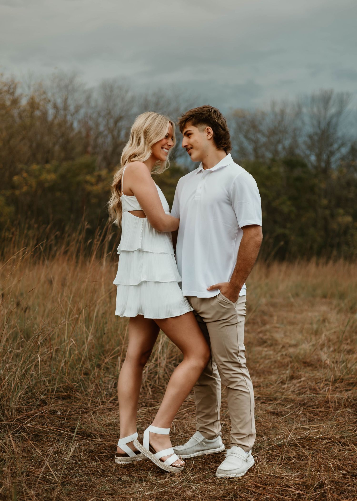 A couple in white outfits share an intimate moment together in a rustic field setting during golden hour.