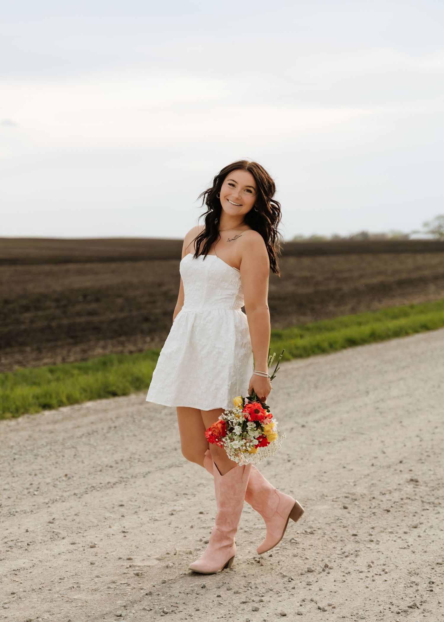 Young woman in white strapless dress and pink cowboy boots holds colorful bouquet on rural dirt road.