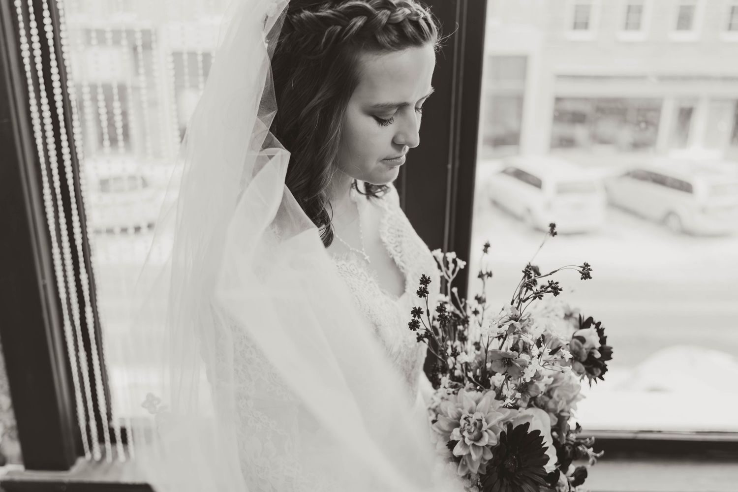 A bride in a veil looks down while holding her bouquet near a window.